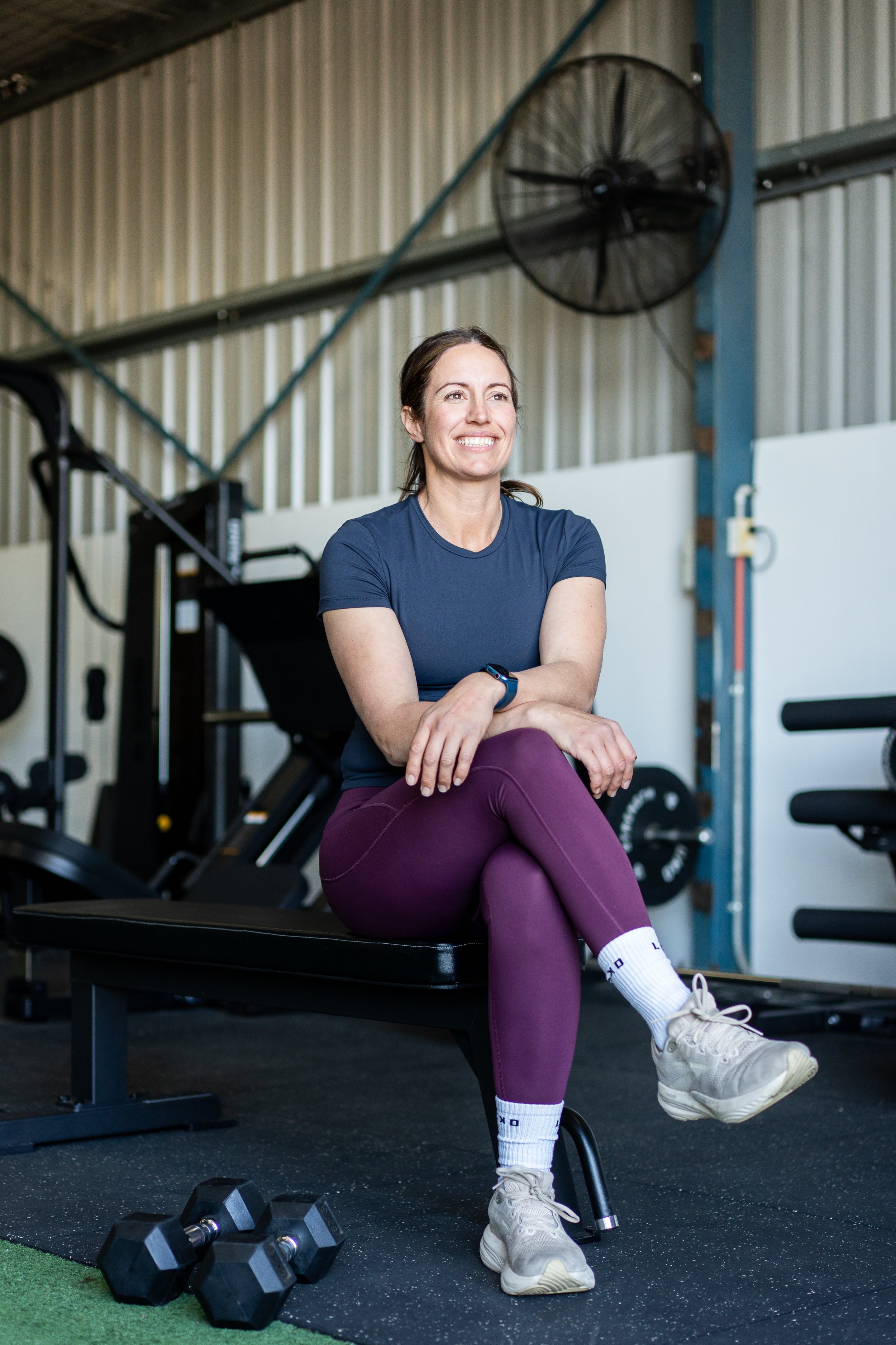 A woman sitting on a workout bench smiling in a gym, wearing a navy T-shirt, purple leggings, white sneakers, and white socks, with dumbbells on the floor nearby.