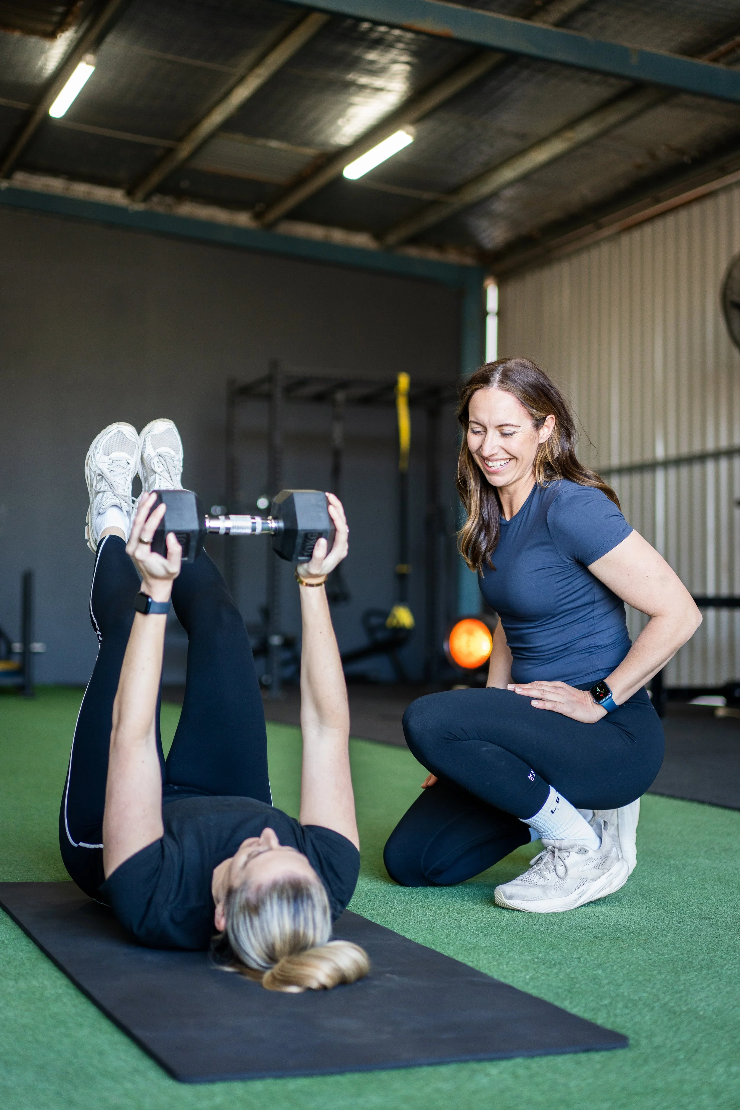 A woman with long brown hair smiling while coaching a woman lying on a black exercise mat; the woman on the mat is lifting dumbbells and has blonde hair.