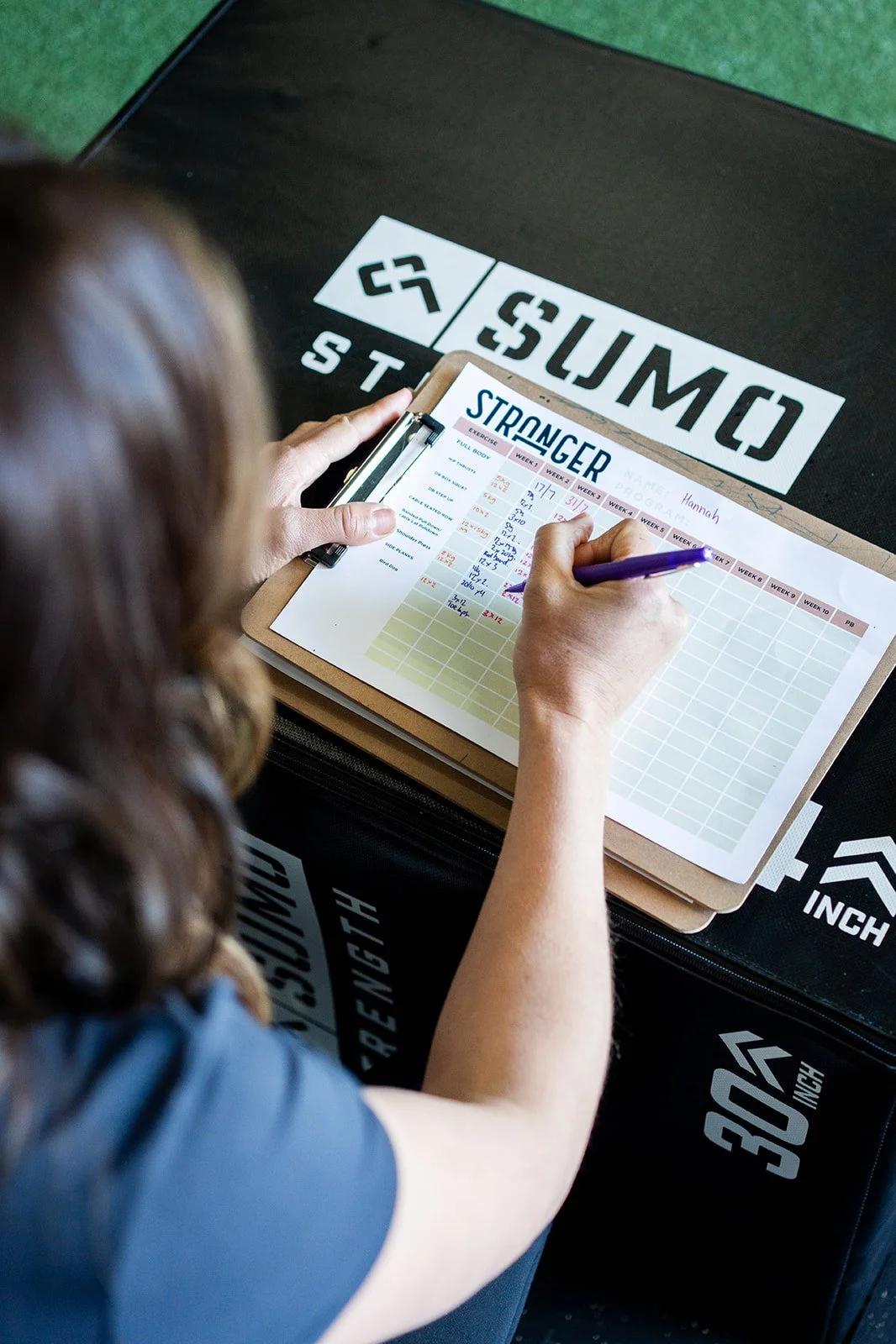 A woman with brown hair is recording her weekly exercise progress on a printed sheet titled 'Stronger' with a purple pen, sitting at a table with workout equipment and stairs in the background.