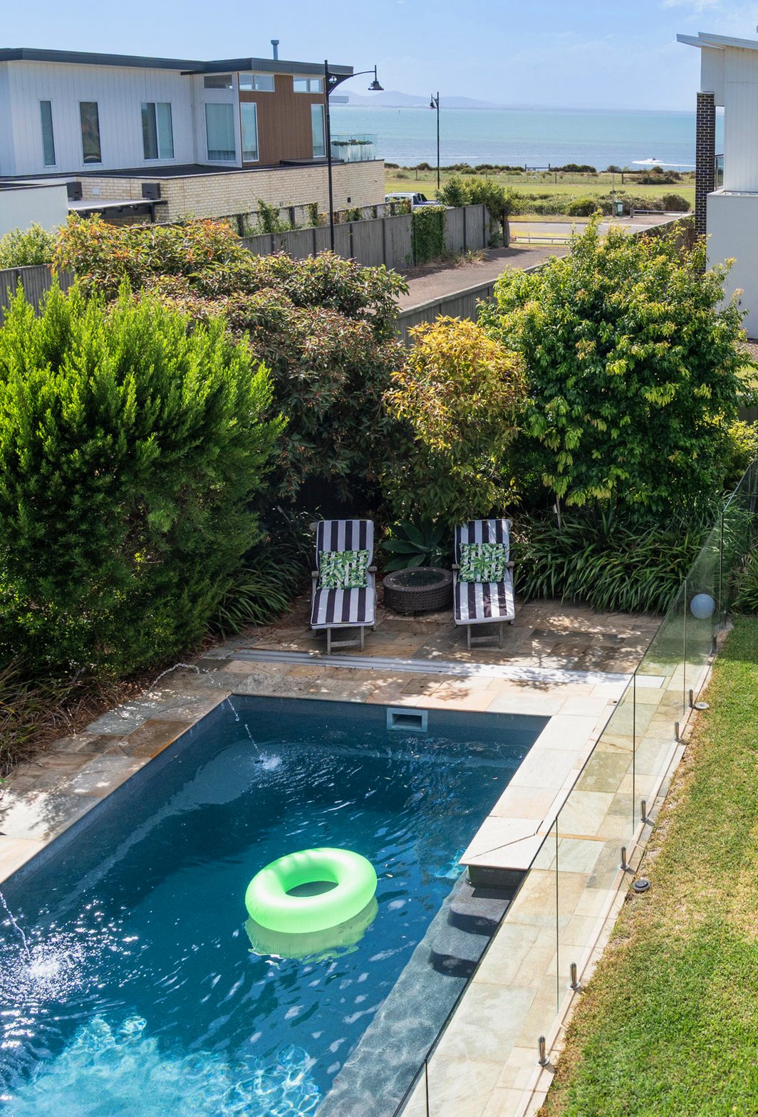 A backyard view featuring a swimming pool with a green float ring, two striped lounge chairs, and lush greenery. A fence separates the yard from a street, with a view of the ocean in the background.