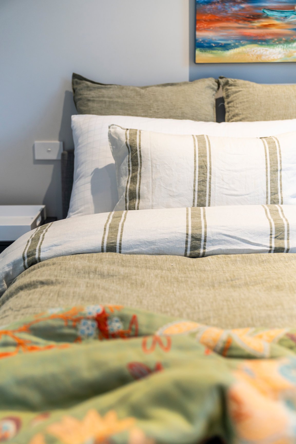 Bed with striped pillows, green blanket, and colorful quilt in a bedroom with a painting on the wall.