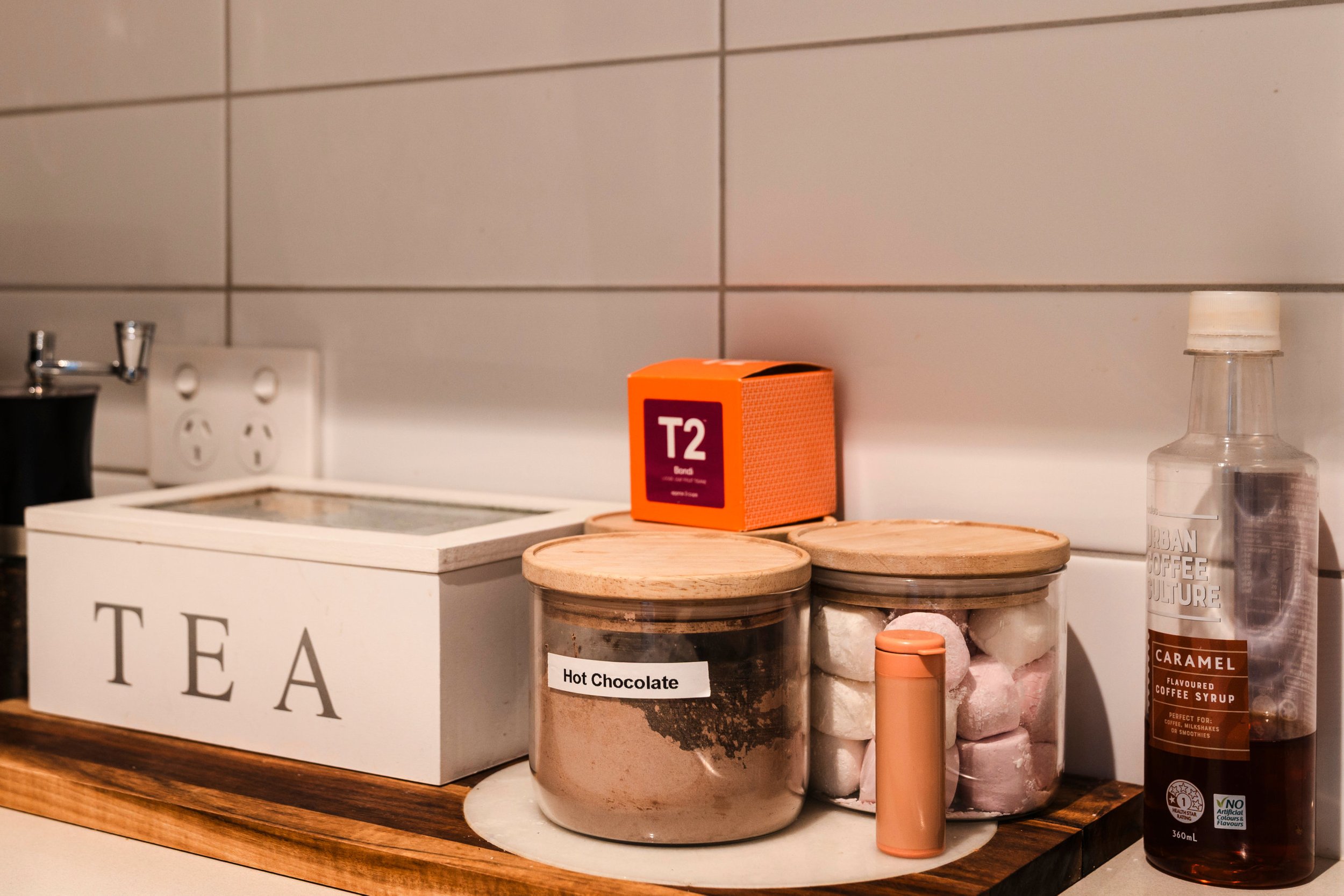 Kitchen counter with a T2 tea box, jars labeled 'Hot Chocolate', marshmallows, caramel syrup bottle, and a white tea box.