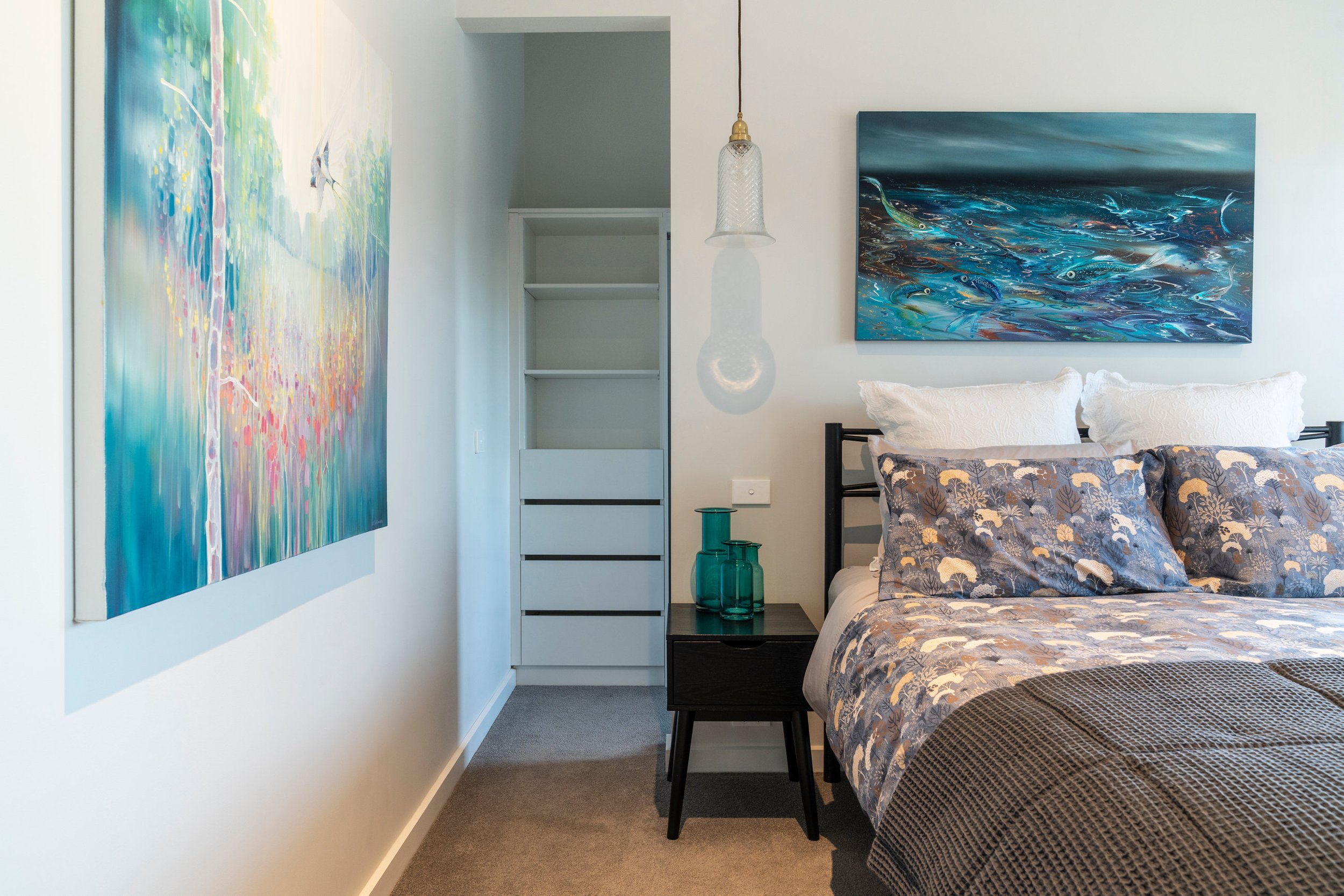 Modern bedroom with a bed featuring patterned bedding, two decorative pillows, a black bedside table with teal glass vases, abstract paintings on the walls, pendant light, and open closet with white shelves and drawers.