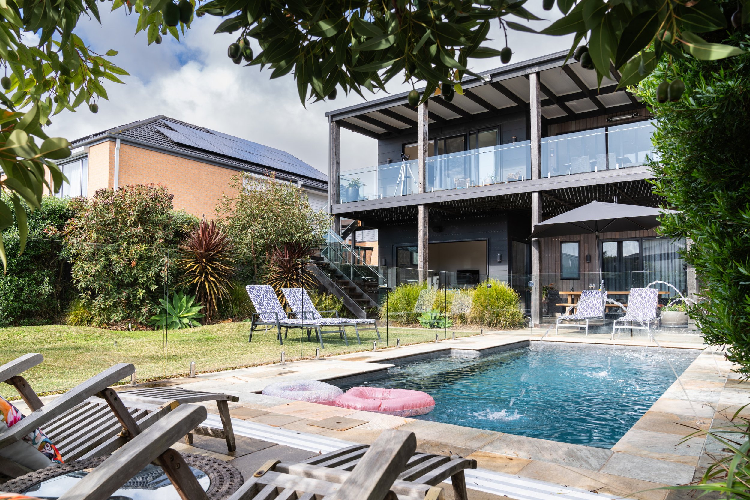 Modern house with solar panels, glass balcony, and a backyard pool with loungers and an umbrella, surrounded by lush greenery.