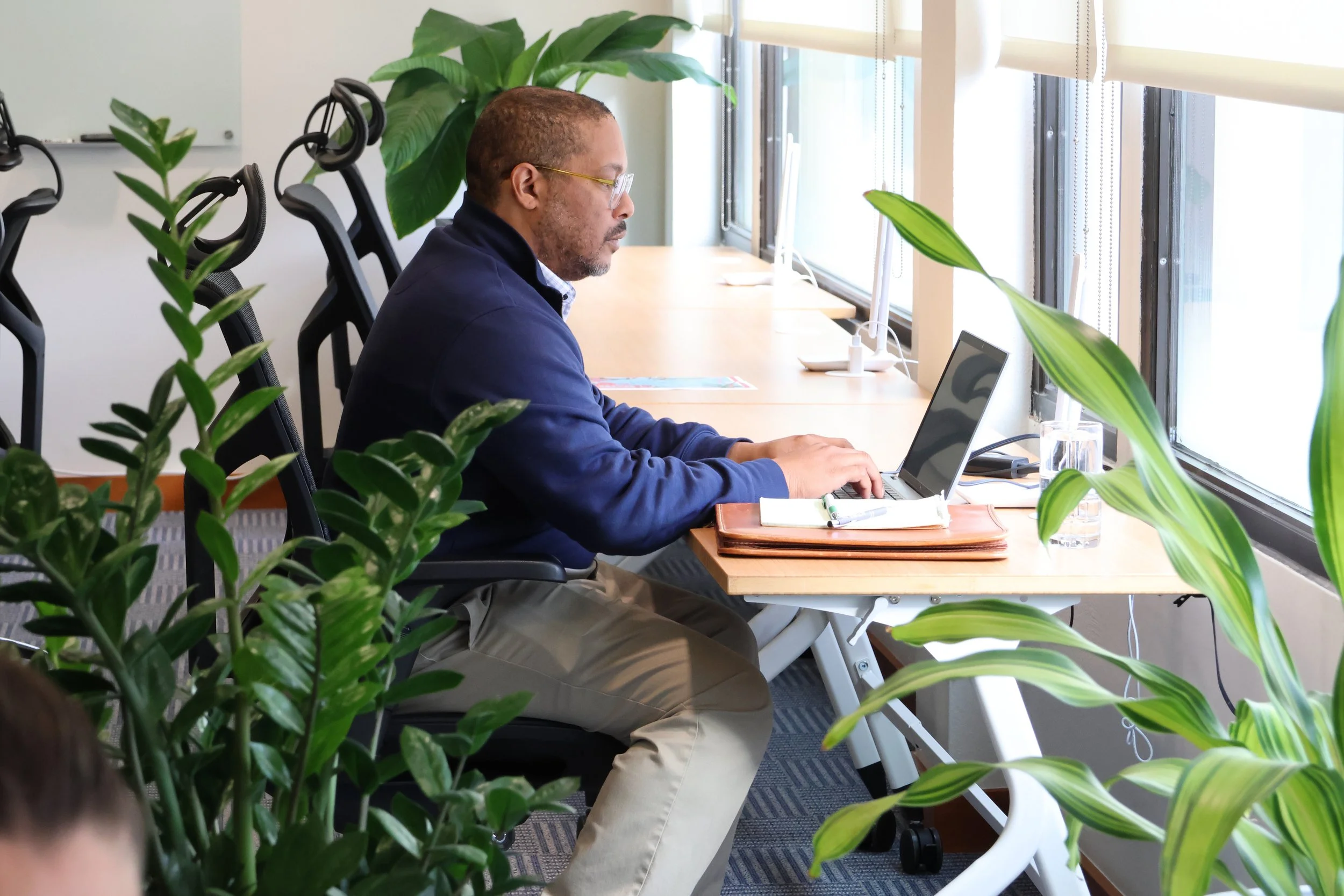 A man with glasses wearing a navy jacket and khaki pants working on a laptop at a desk near a window.