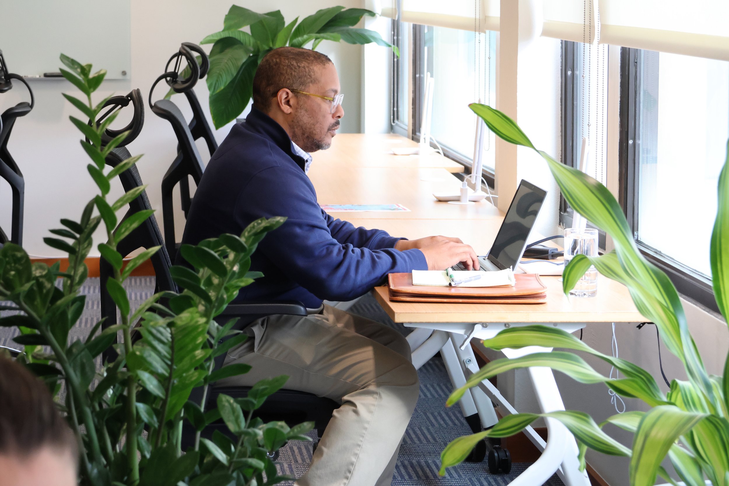 A man sitting at a desk working on a laptop in an office with large windows and  plants.