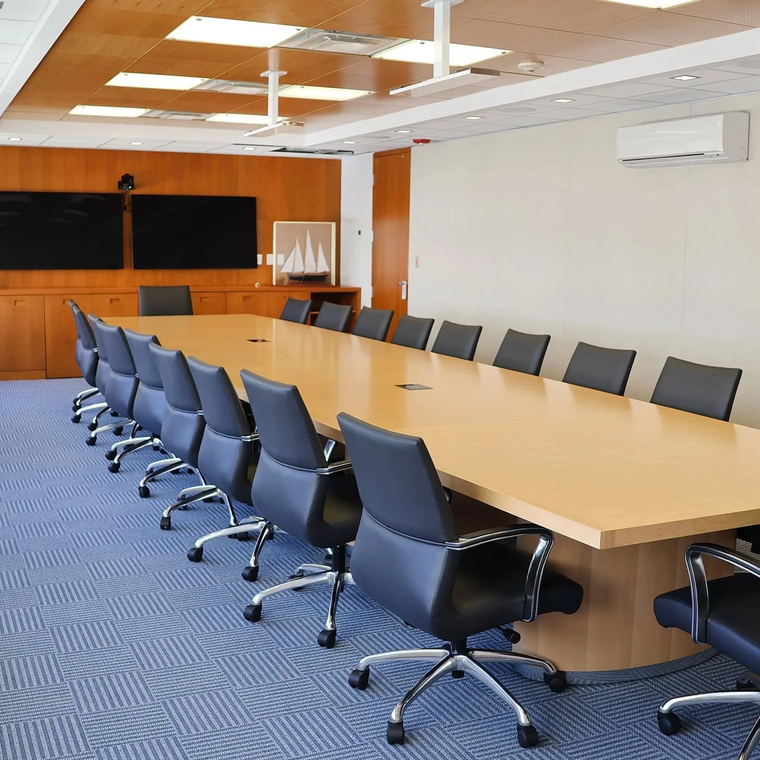 Conference room with a long wooden table surrounded by black swivel chairs, wooden panel walls, a blue carpet, and two large screens on the back wall.