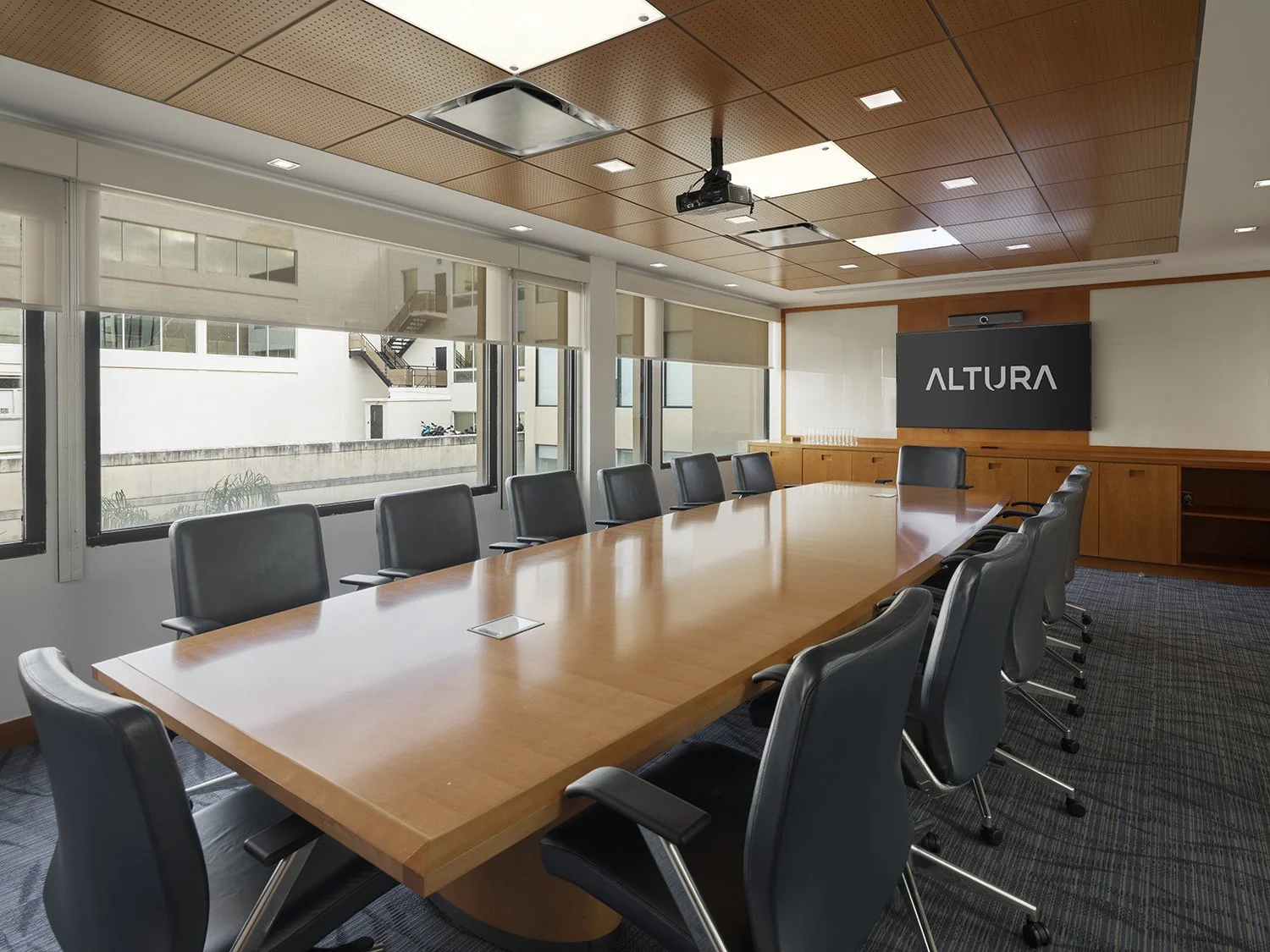 Conference room with a long wooden table, black leather chairs, projector, and large screen on the wall displaying "ALTURA" text.