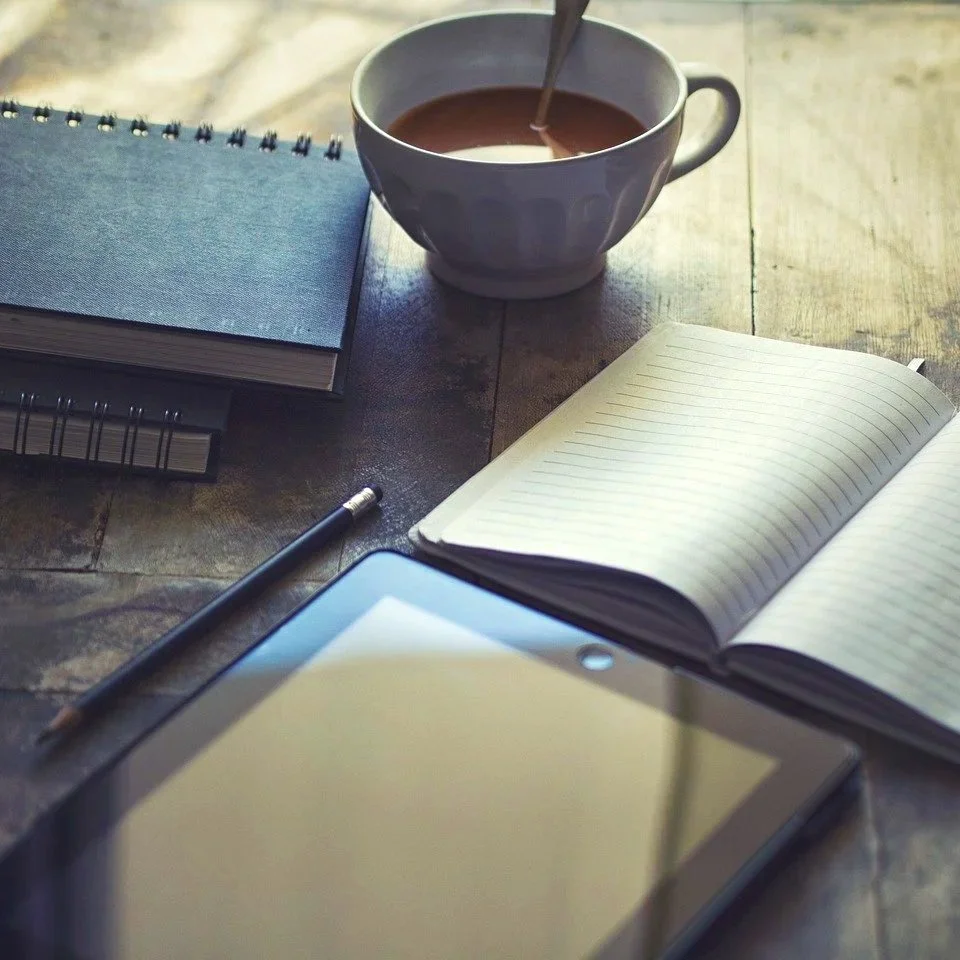 A cup of coffee, notebooks, a pen, an open graph paper notebook, and a tablet on a wooden table.