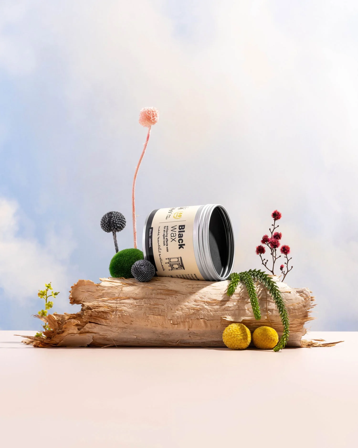 A container labeled "Black Wax" resting on a piece of bark, surrounded by various small, colorful, decorative plants and flowers against a soft, sky-like background.