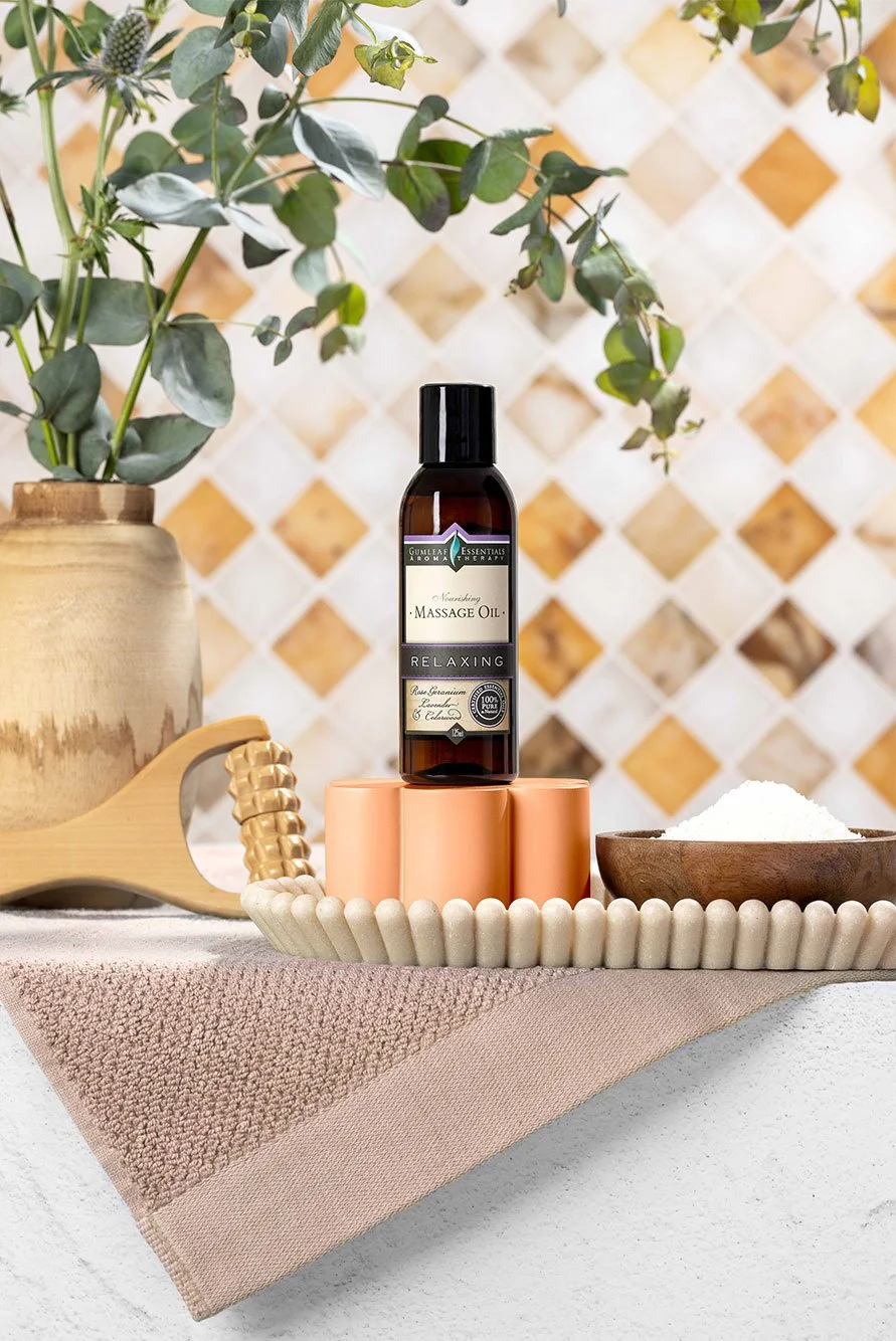 Spa setting with massage oil, wooden massager, ornamental vase with greenery, and bowl of bath salts on beige towel.