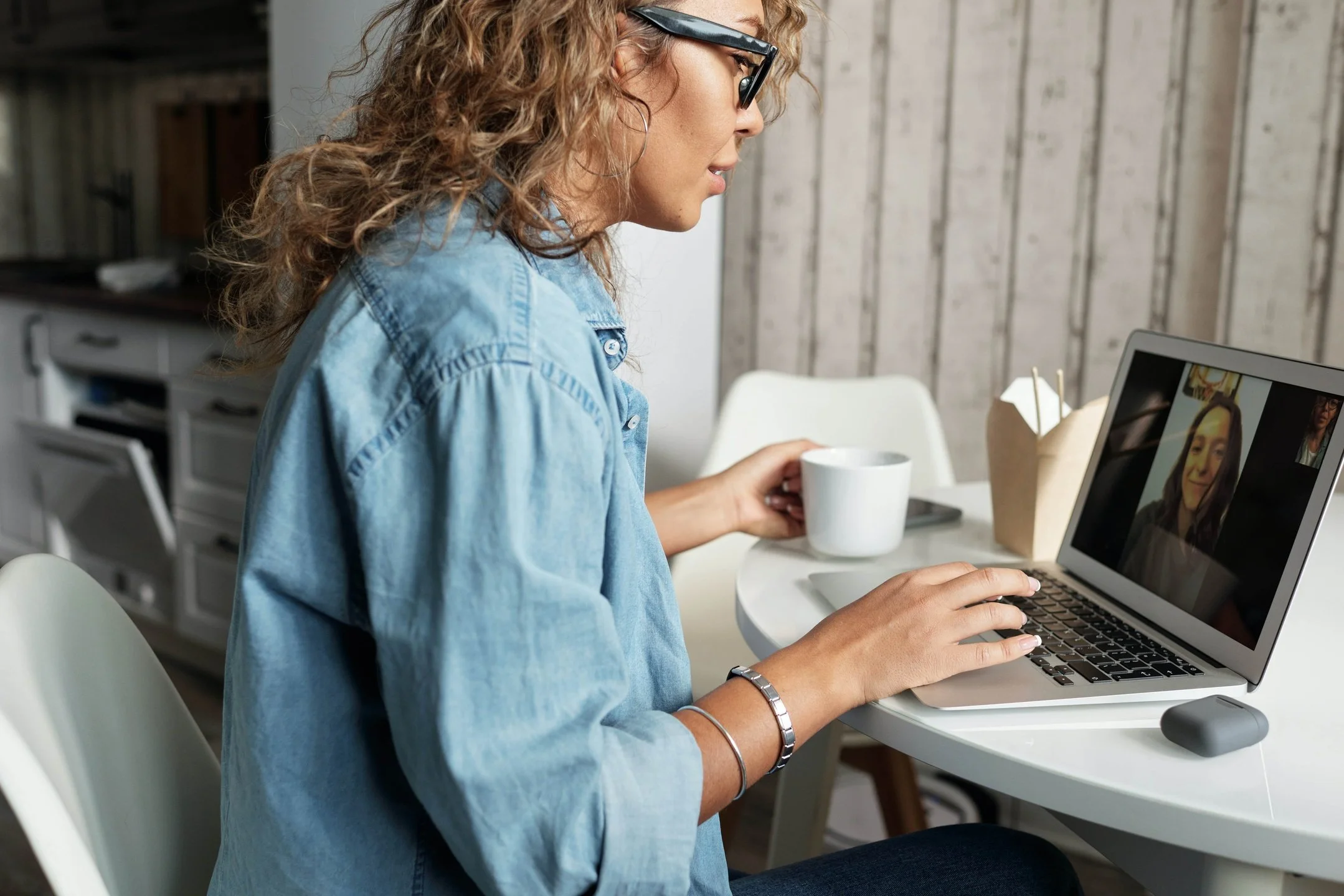 Woman with curly hair and glasses in a denim shirt sitting at a table, holding a white mug, and using a laptop for a video call in a cozy kitchen or dining area.
