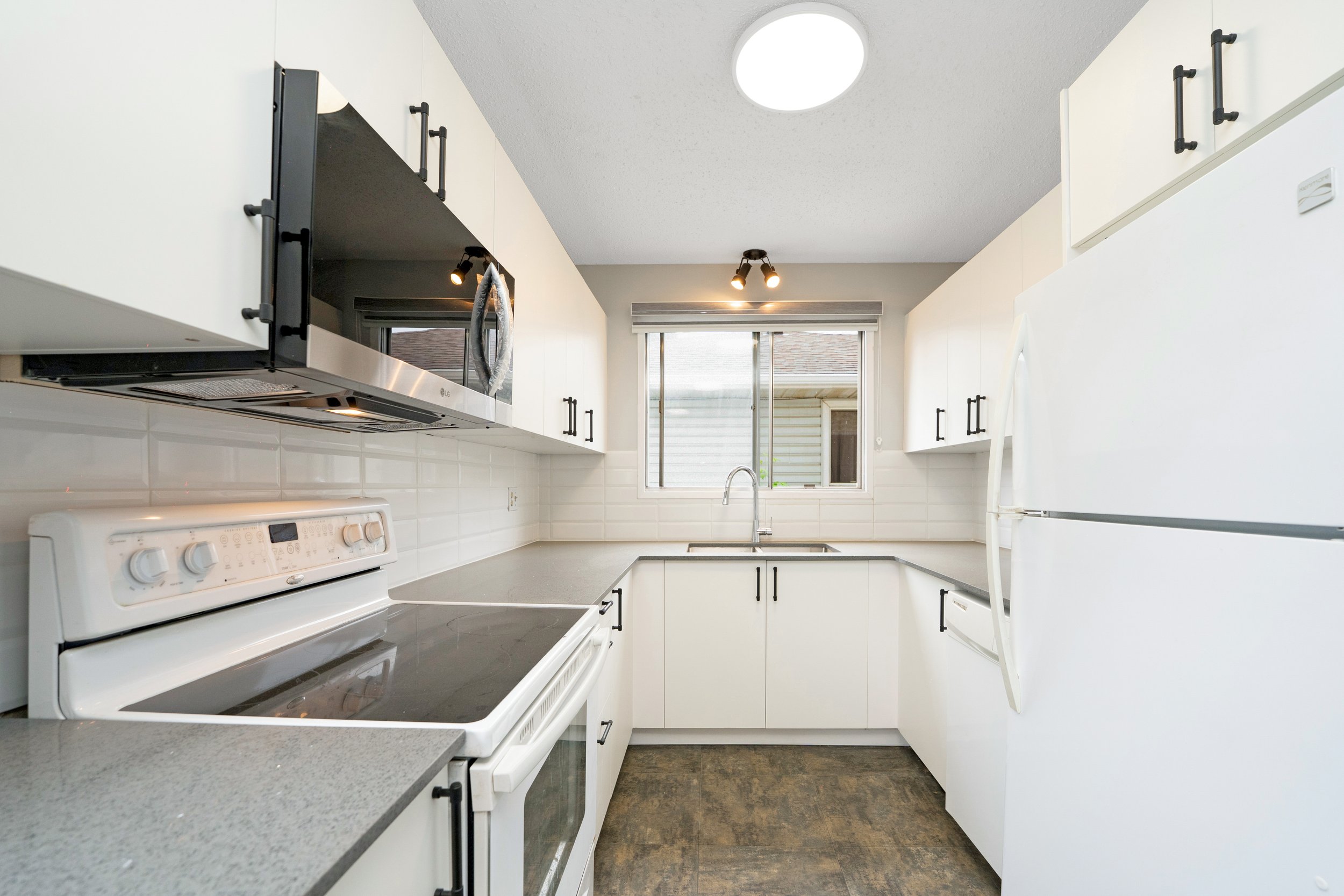 White kitchen with black handles, microwave, oven, refrigerator, white subway tile backsplash, and a window above the sink.
