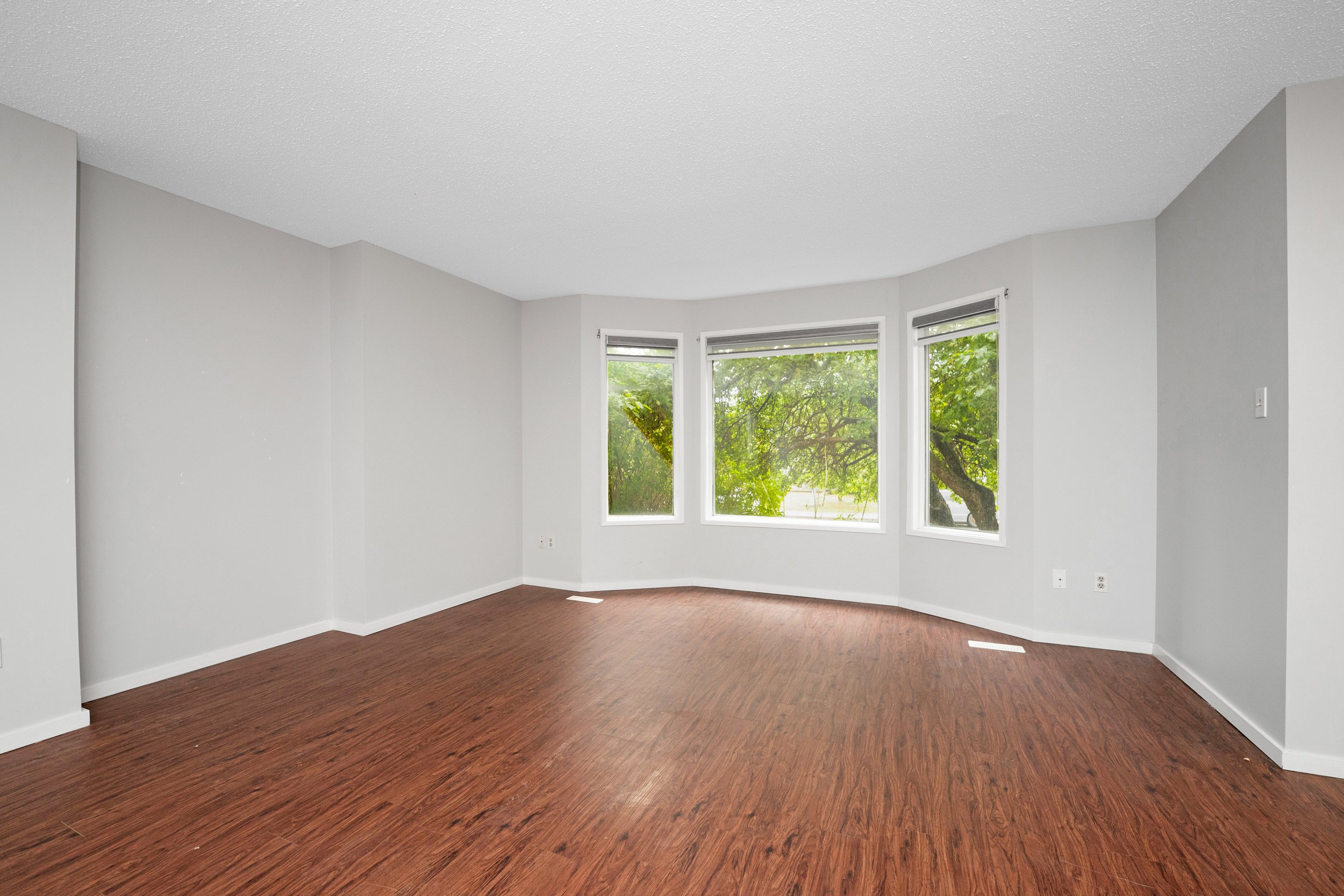 Empty living room with light gray walls, wooden floor, and large windows with green trees visible outside.