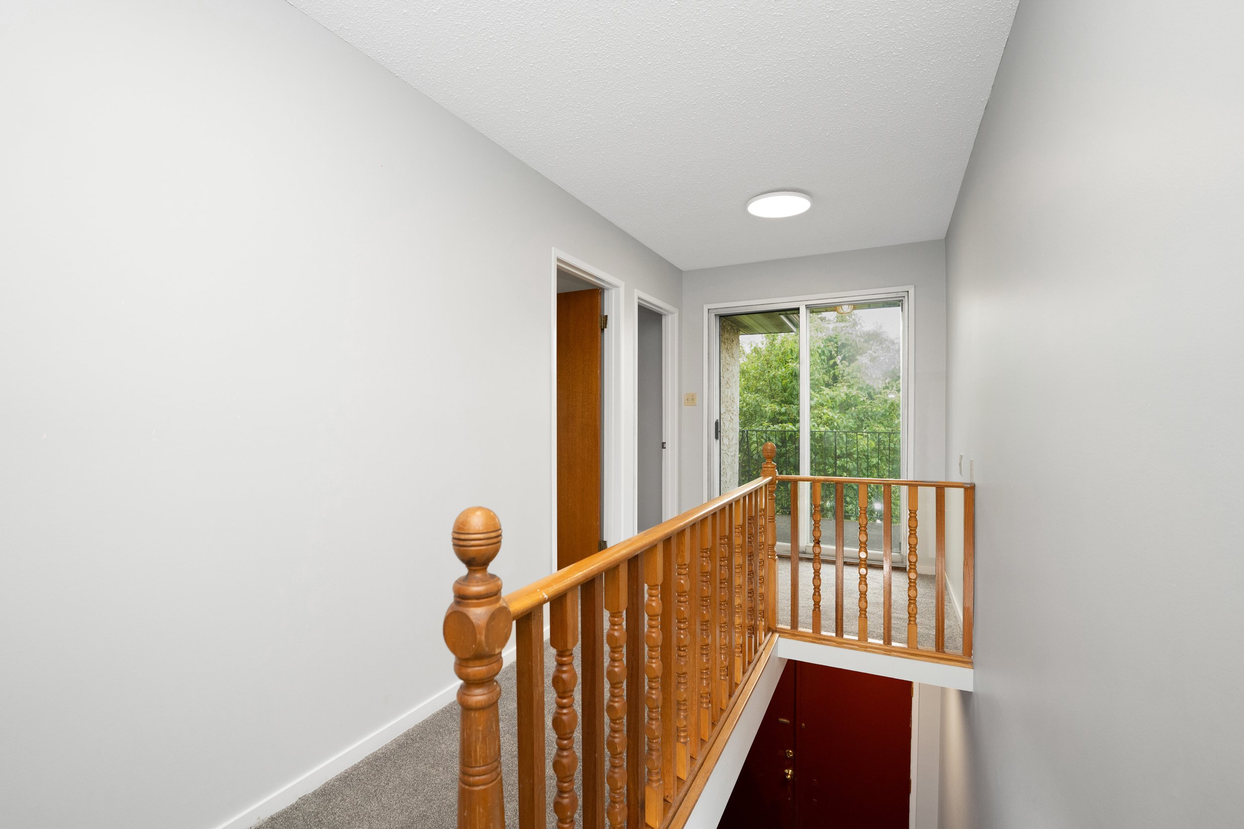 Interior of an upstairs hallway with a wooden railing, a sliding glass door leading to a balcony, white walls, carpeted flooring, and ceiling light fixture.