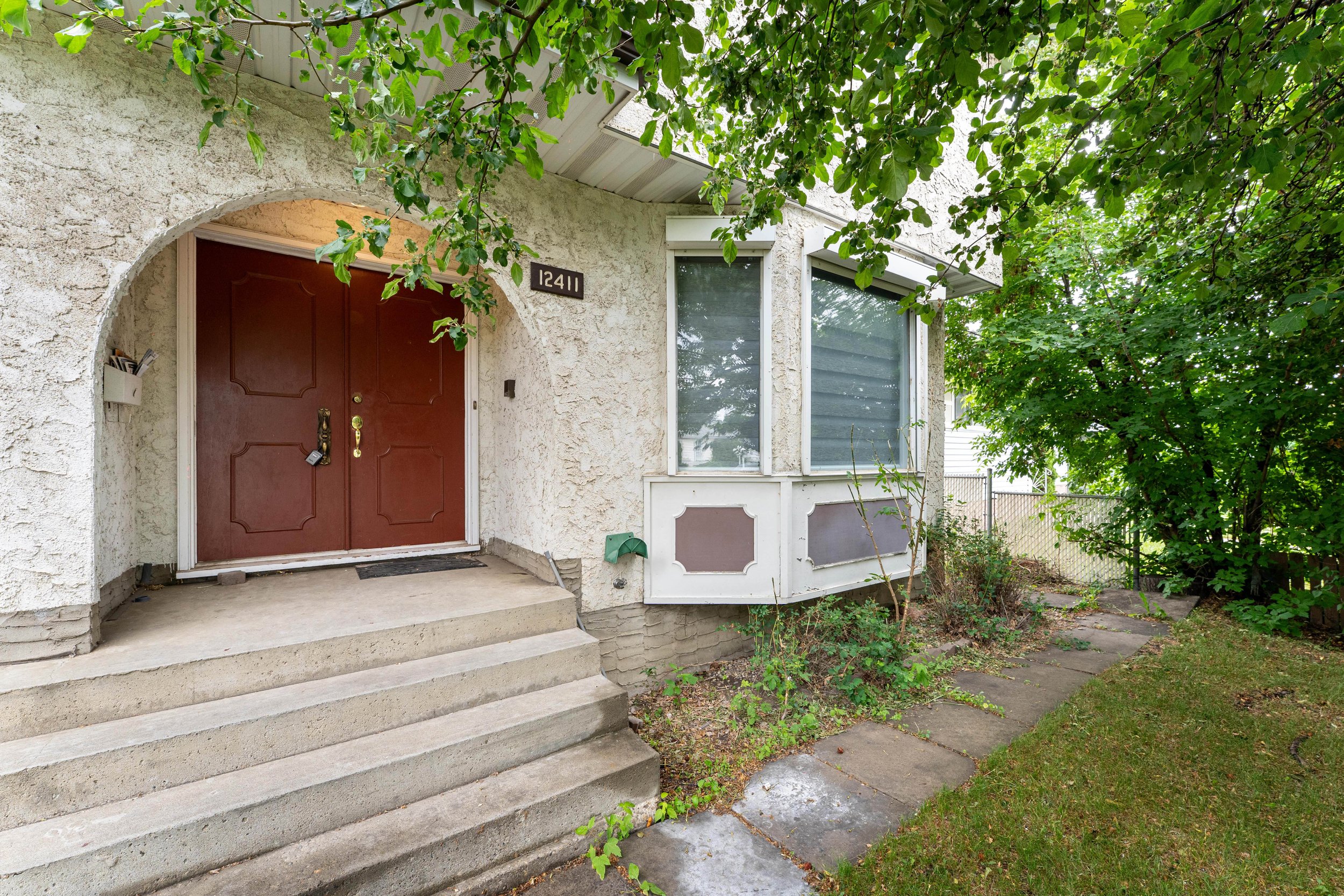 Front entrance of a house with red double doors, concrete steps, white stucco exterior, and overgrown bushes on a cloudy day.