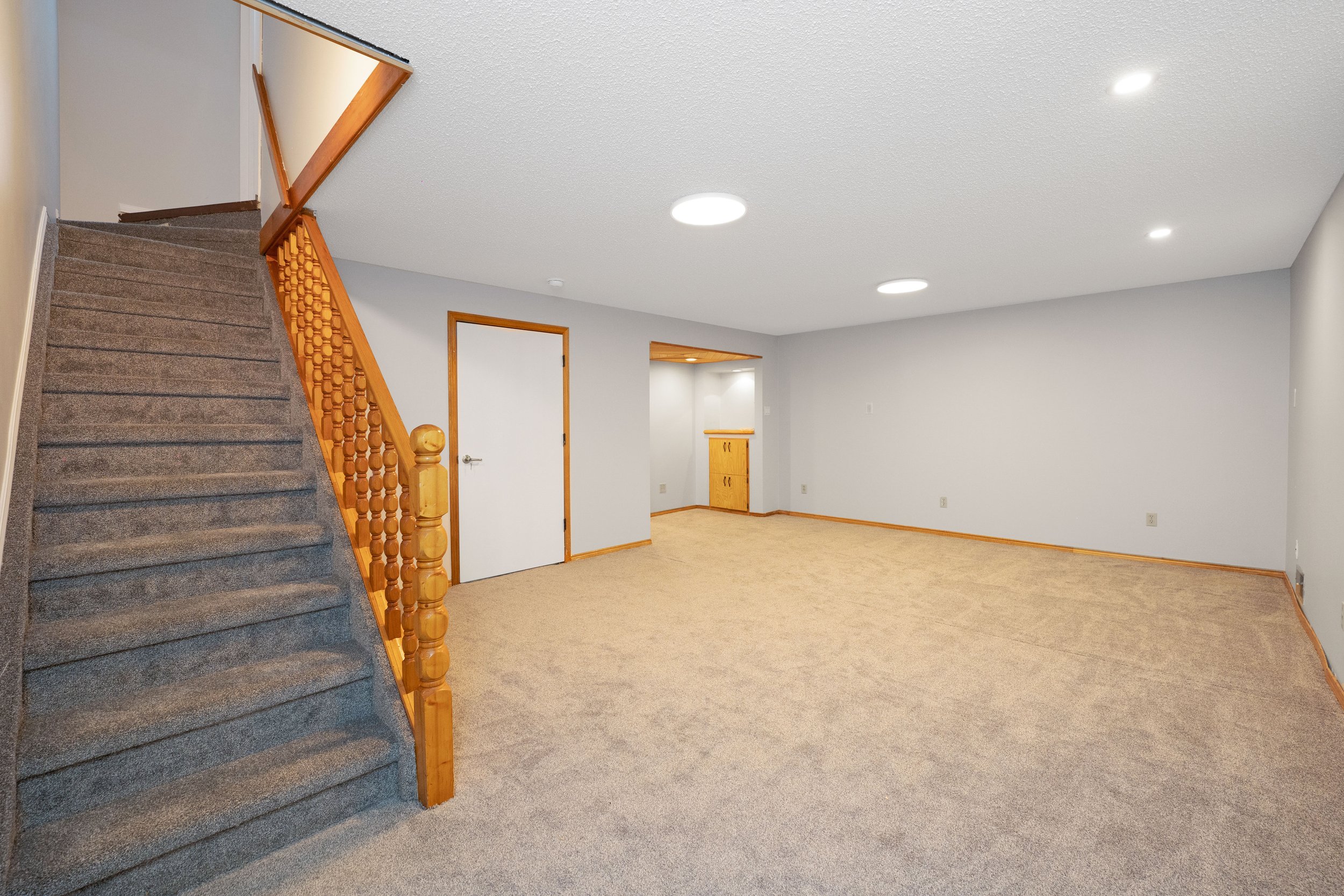 Empty living room with beige carpet, white walls, wood trim, staircase with gray carpet, built-in wooden cabinet in corner, and ceiling lights.