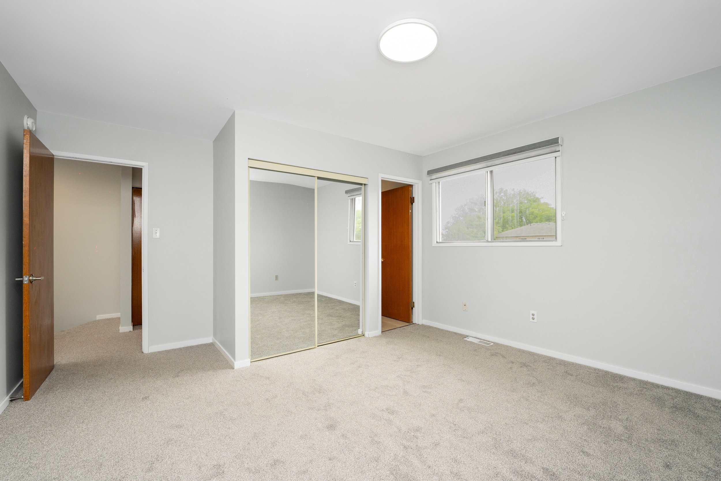 Empty bedroom with beige carpet, white walls, a closet with mirrored sliding doors, a window, a wooden door, and a ceiling light