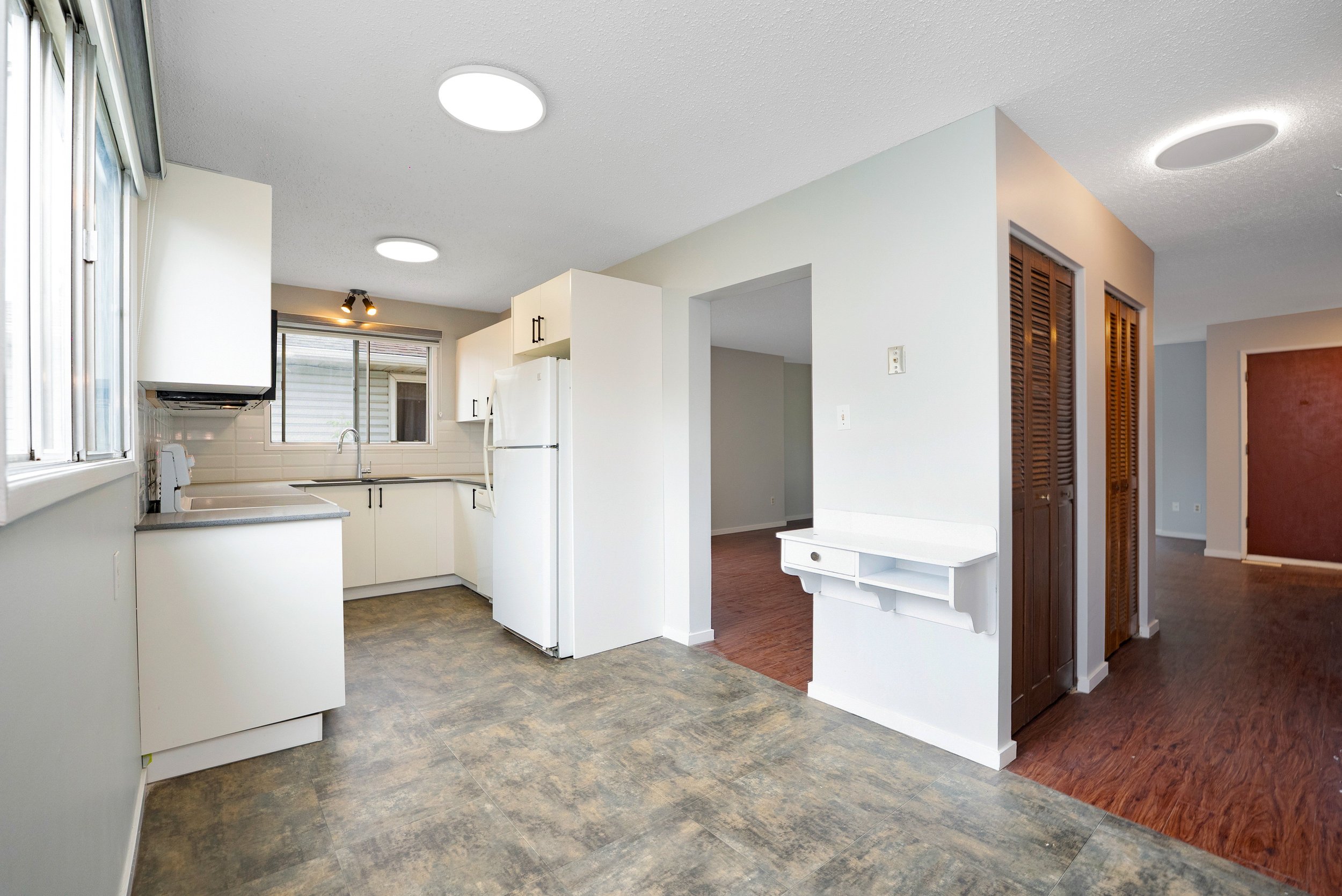 Empty kitchen with white cabinets, white refrigerator, and windowed view outside. Adjacent to a hallway with wooden floors and dark brown louvered closet doors.