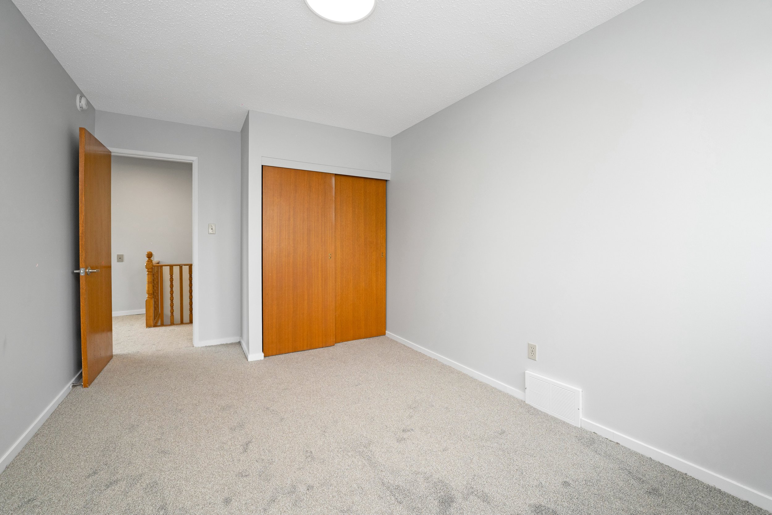Empty bedroom with light gray walls, beige carpet, wooden closet doors, and door open to hallway showing staircase.