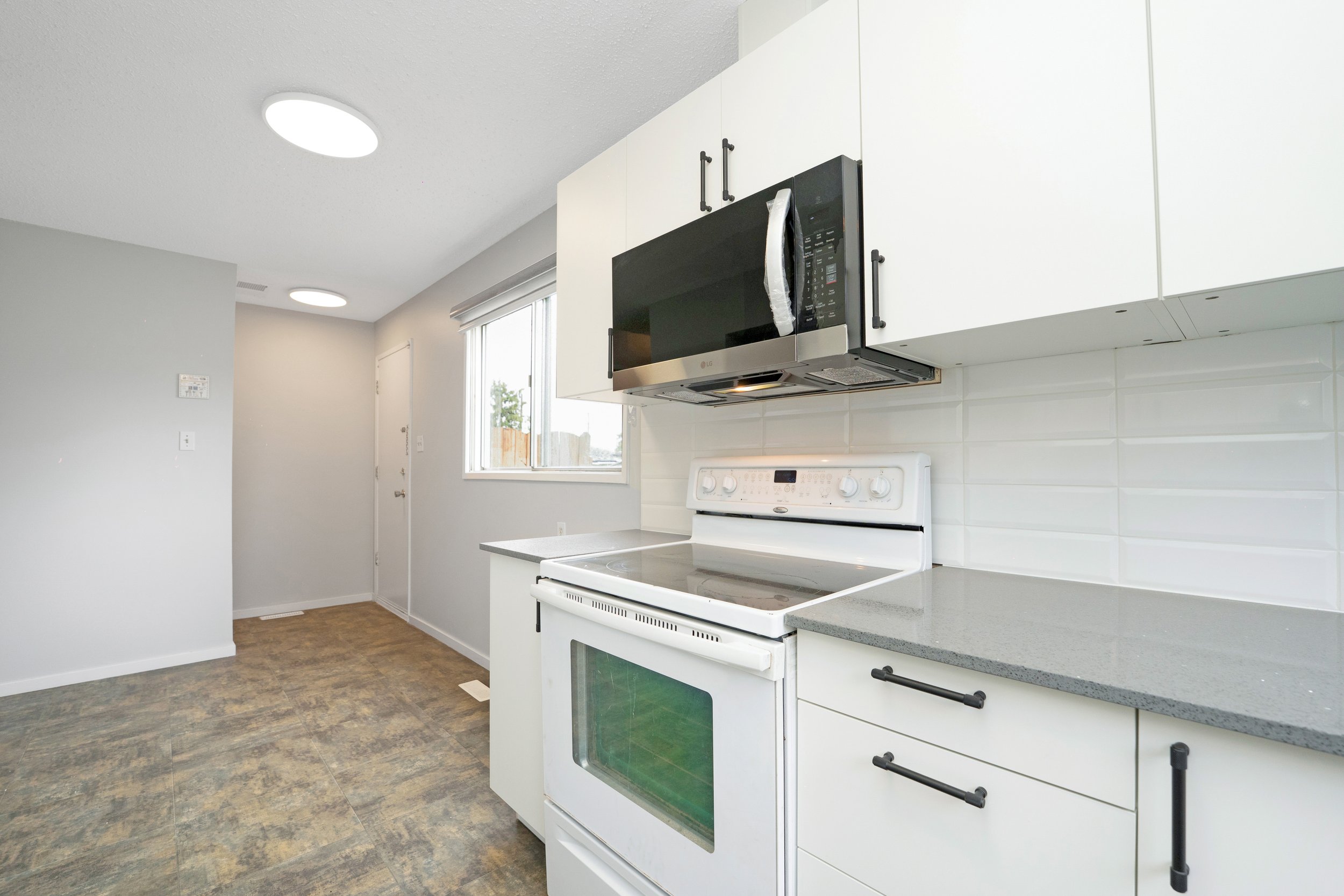 Modern kitchen with white cabinets, gray countertop, microwave, white stove, and a window.