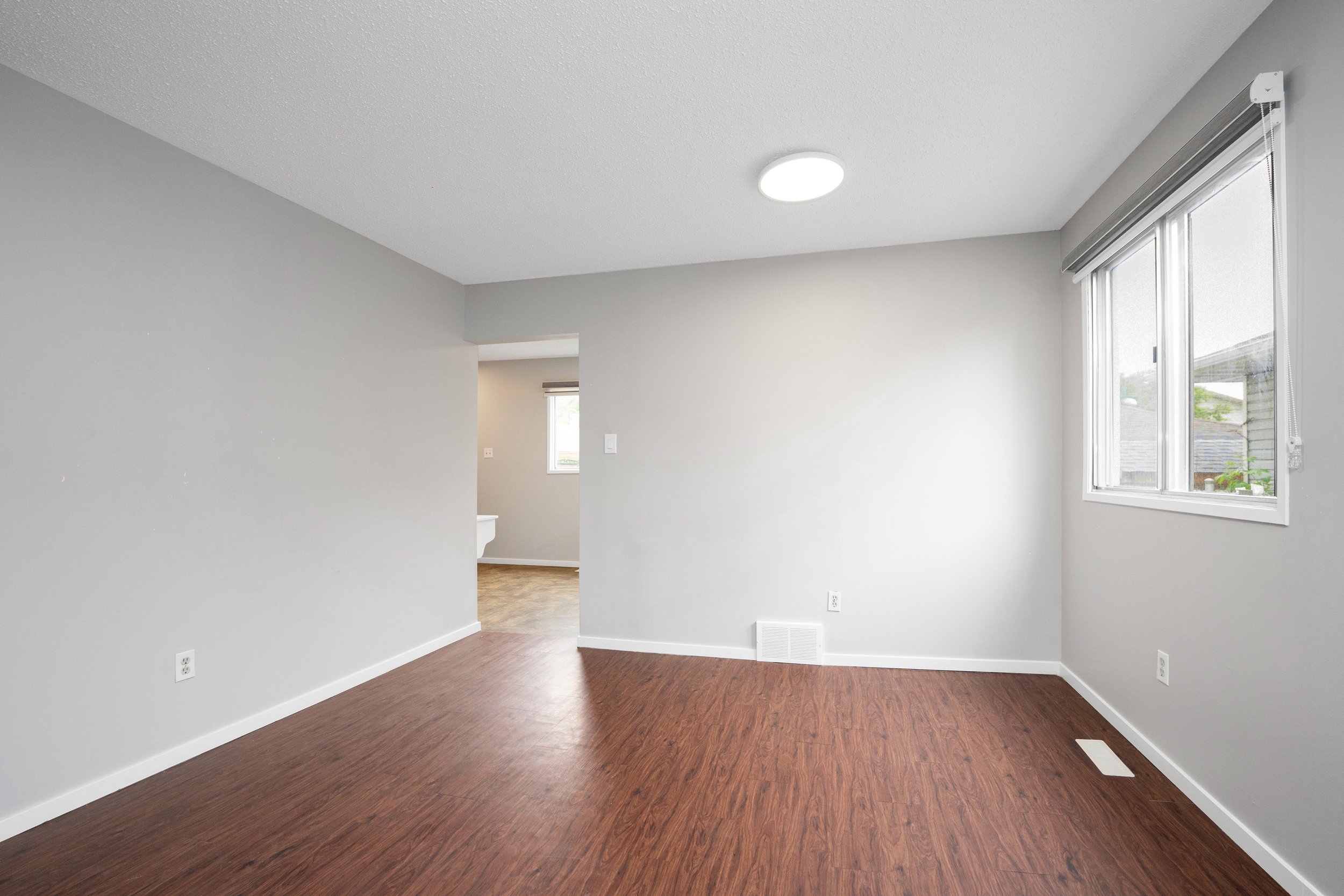 Empty room with gray walls, hardwood floors, and a window with blinds, with a view into a bathroom in the background.