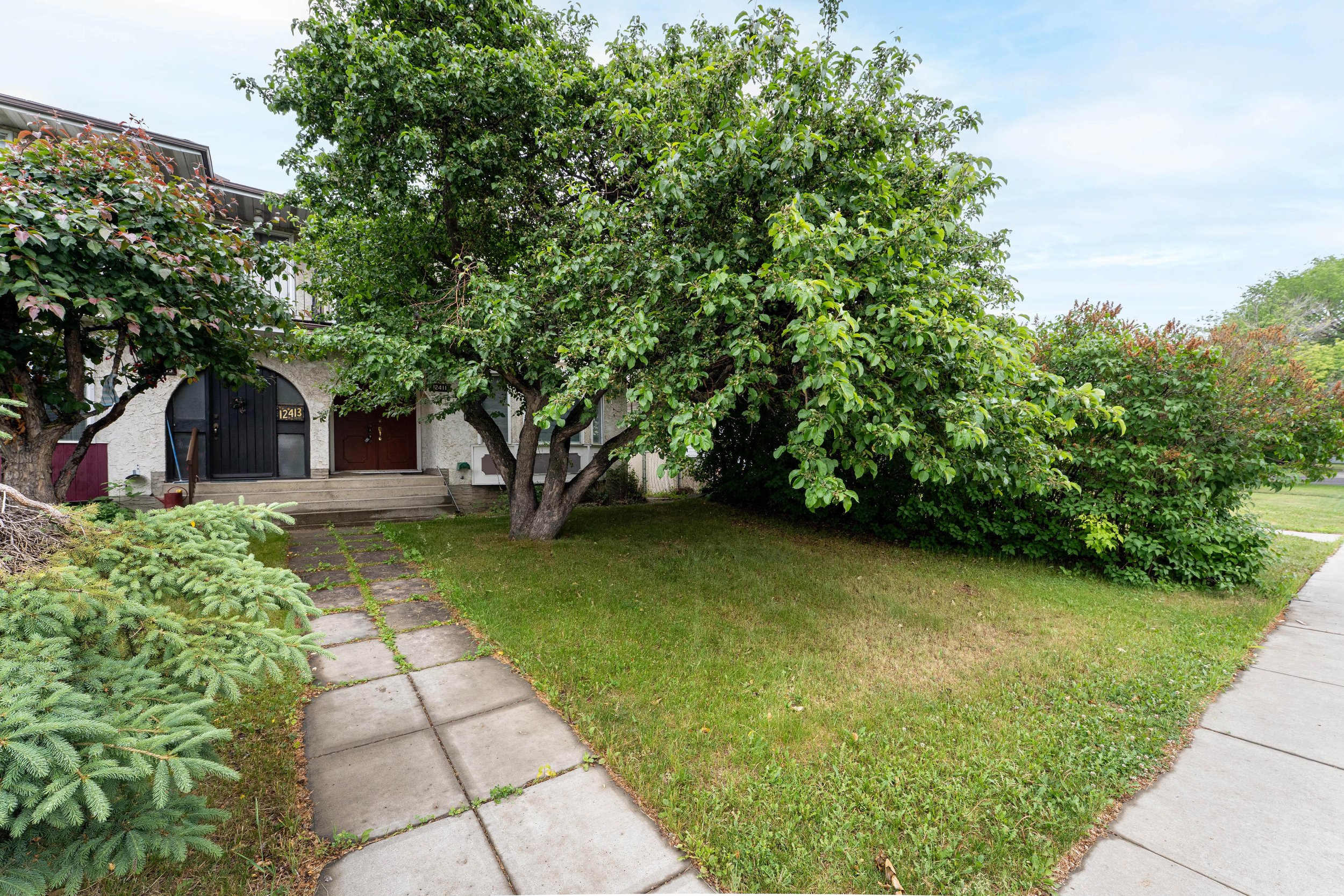 Front yard of a house with a sidewalk, green grass, trees, and shrubbery in a suburban neighborhood.