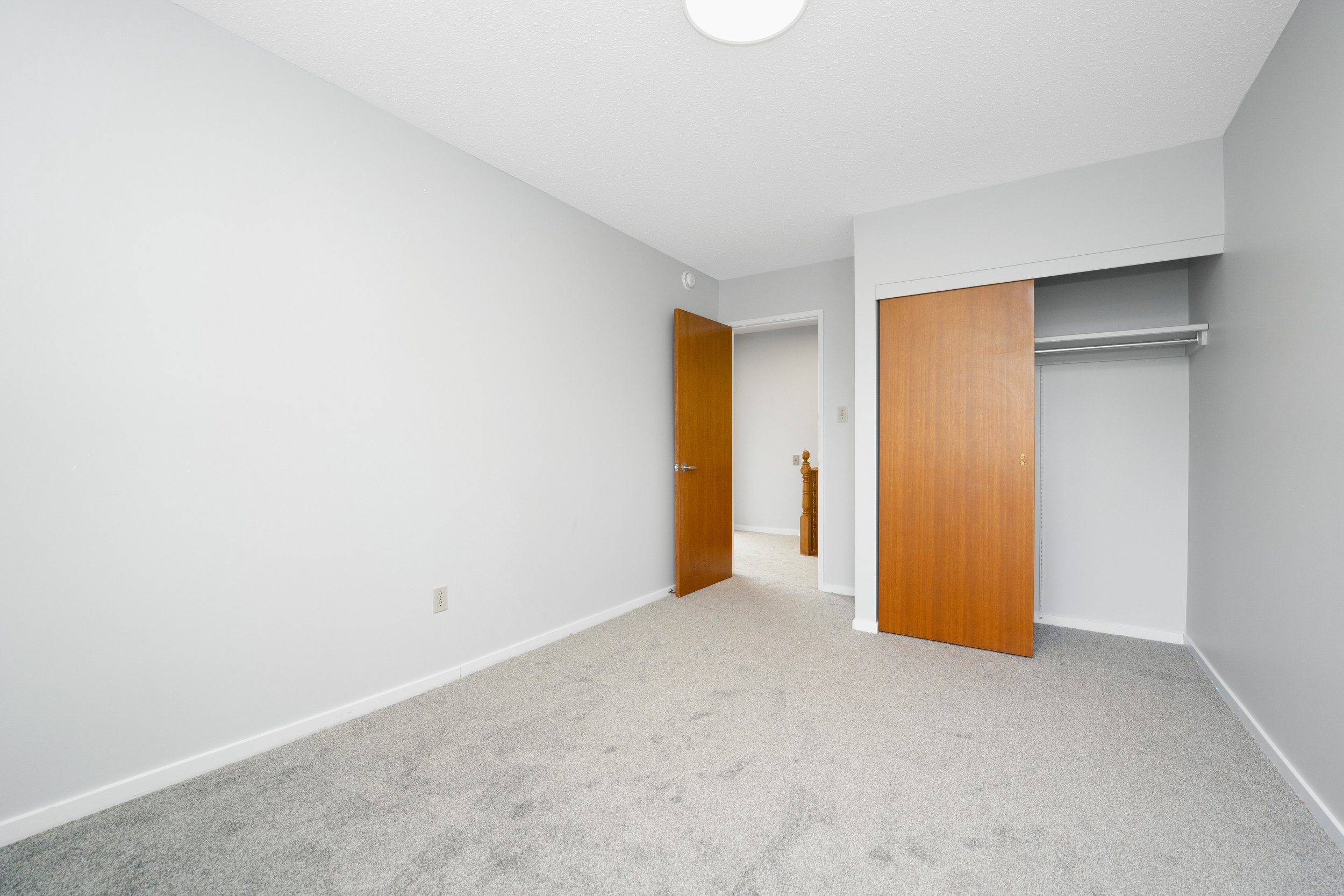 Empty bedroom with light gray walls, beige carpet, a closet with wooden sliding doors, and a partially open wooden door leading to a hallway.