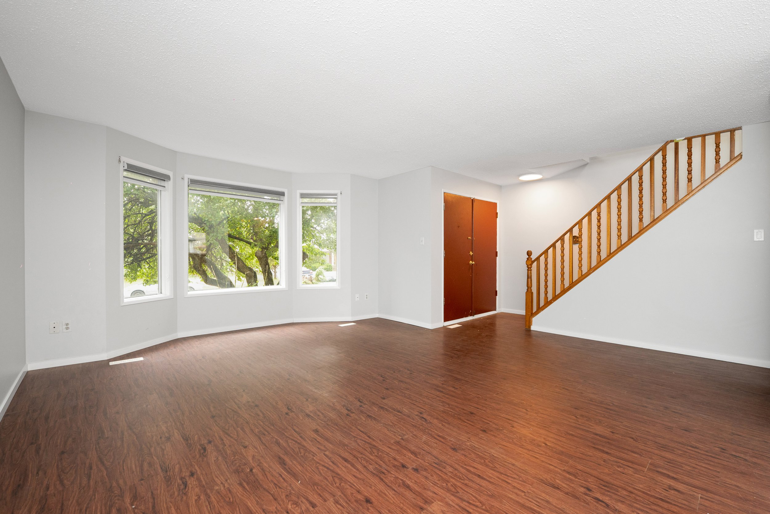 Empty living room with three large windows, a red front door, wooden flooring, and a staircase with a wooden railing leading upstairs.