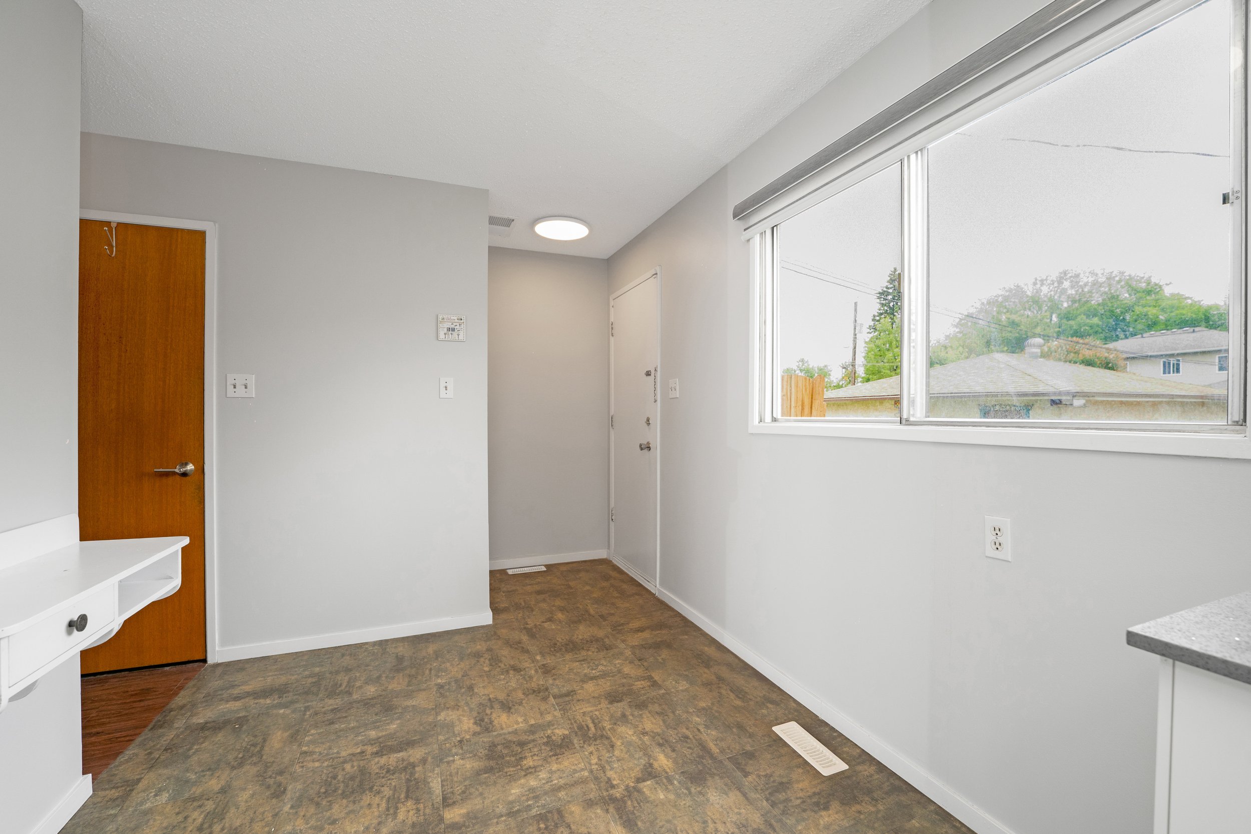 Empty kitchen area with a white wall, a brown door, a window with a view of a neighborhood, and a brown floor.