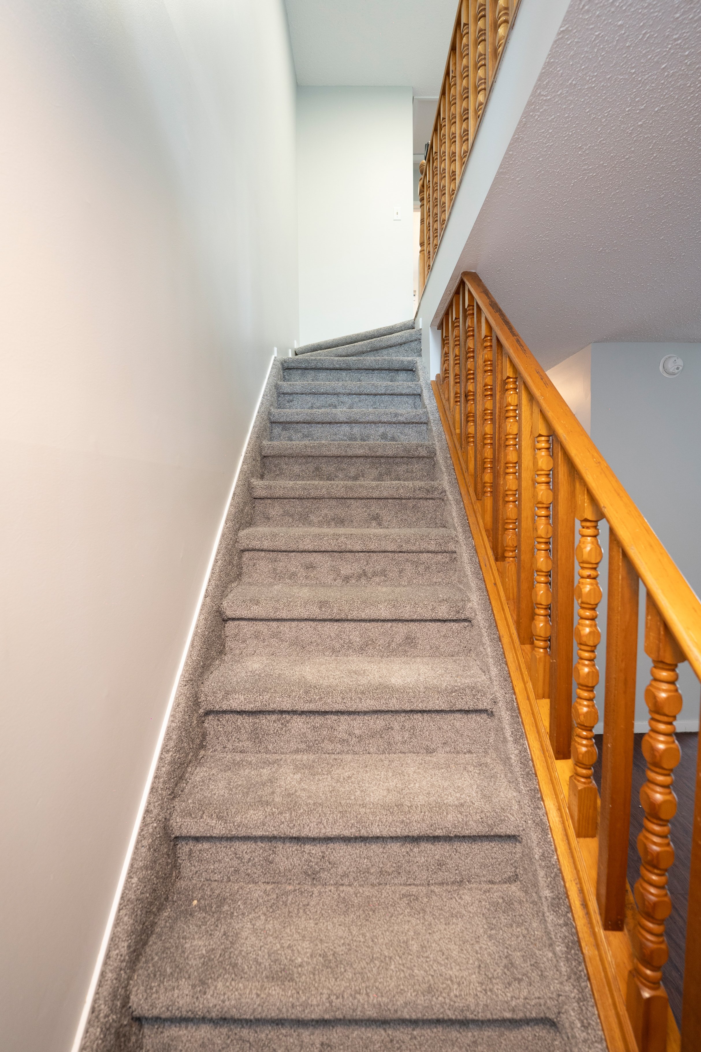 Image of a carpeted staircase inside a house, with a wooden railing on the right side and a plain wall on the left side, leading to an upper floor with white walls.