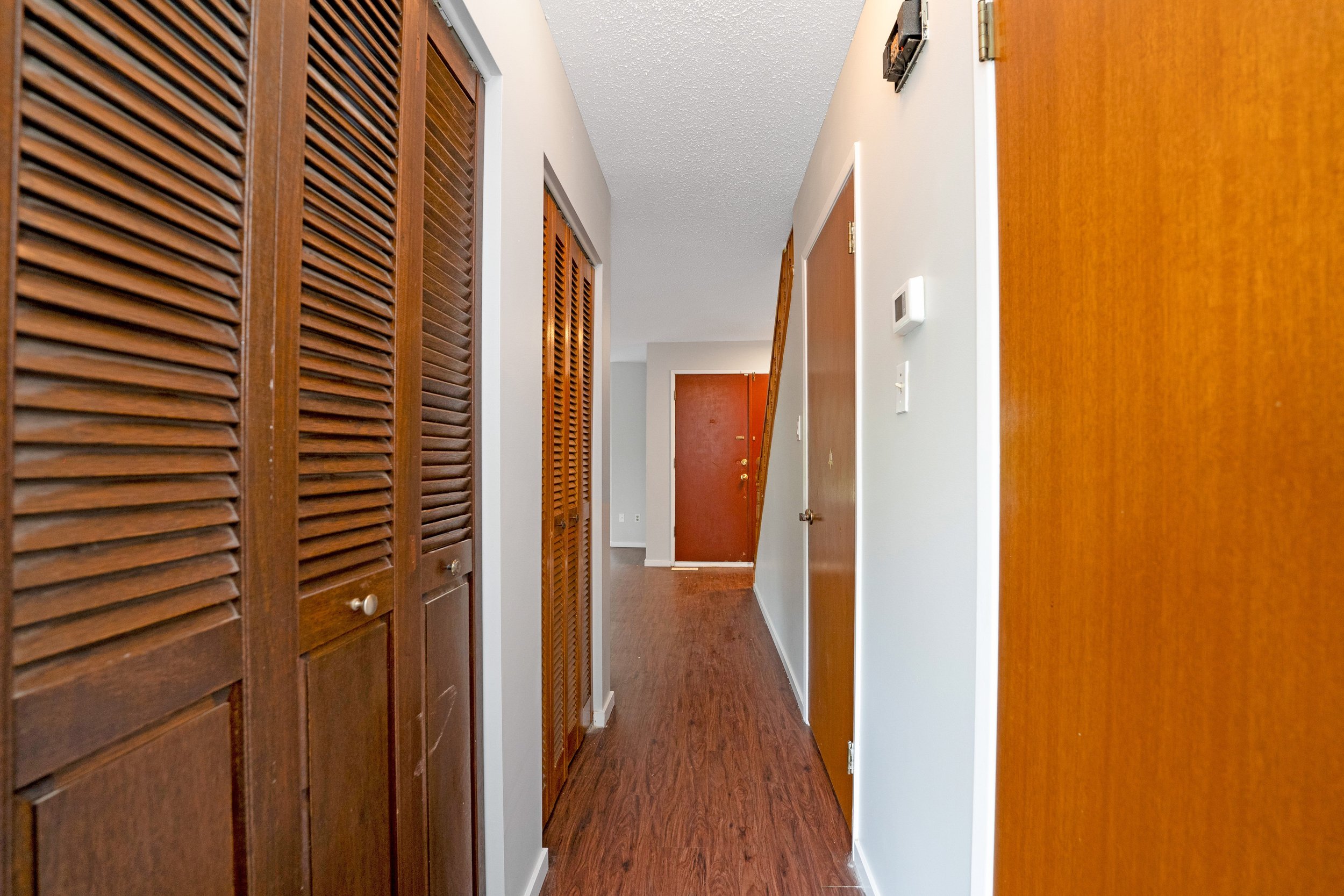 Hallway with brown wooden doors and white walls, hardwood floor, and a front door at the end.