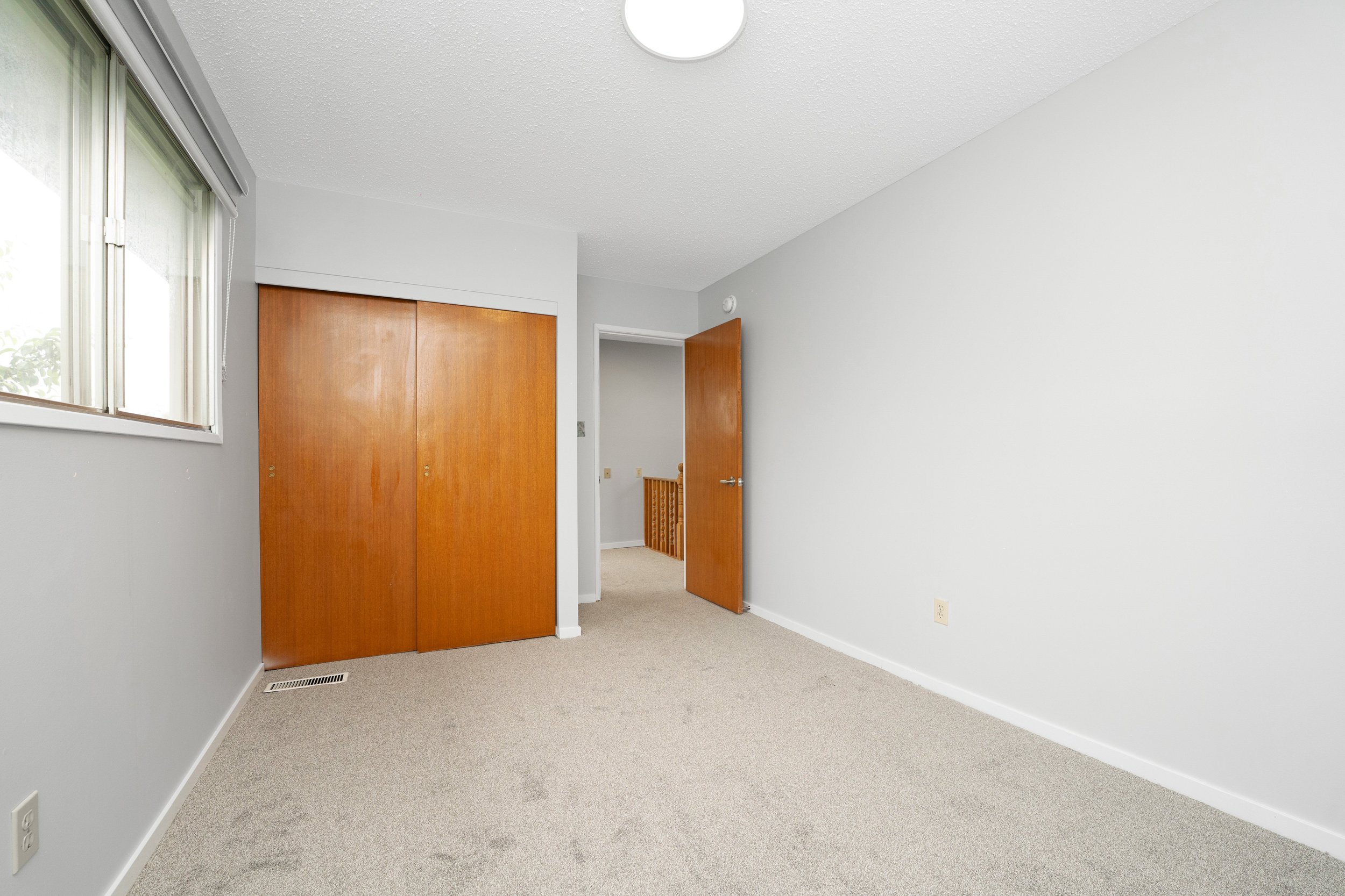 Empty bedroom with beige carpet, white walls, a window with blinds, a wooden closet door, and an open door leading to a hallway with a wooden staircase railing.