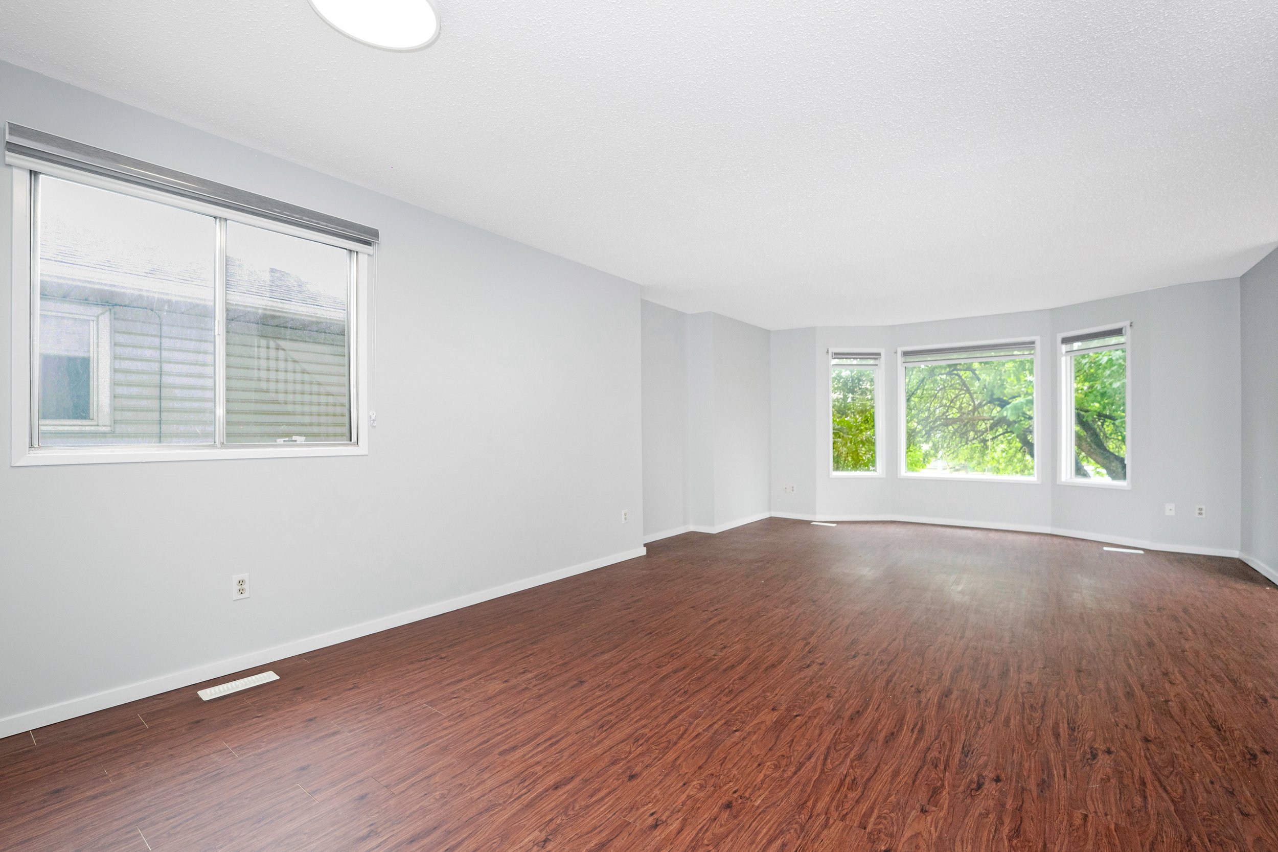 Empty living room with white walls, large windows, and wooden flooring, overlooking a green tree outside.