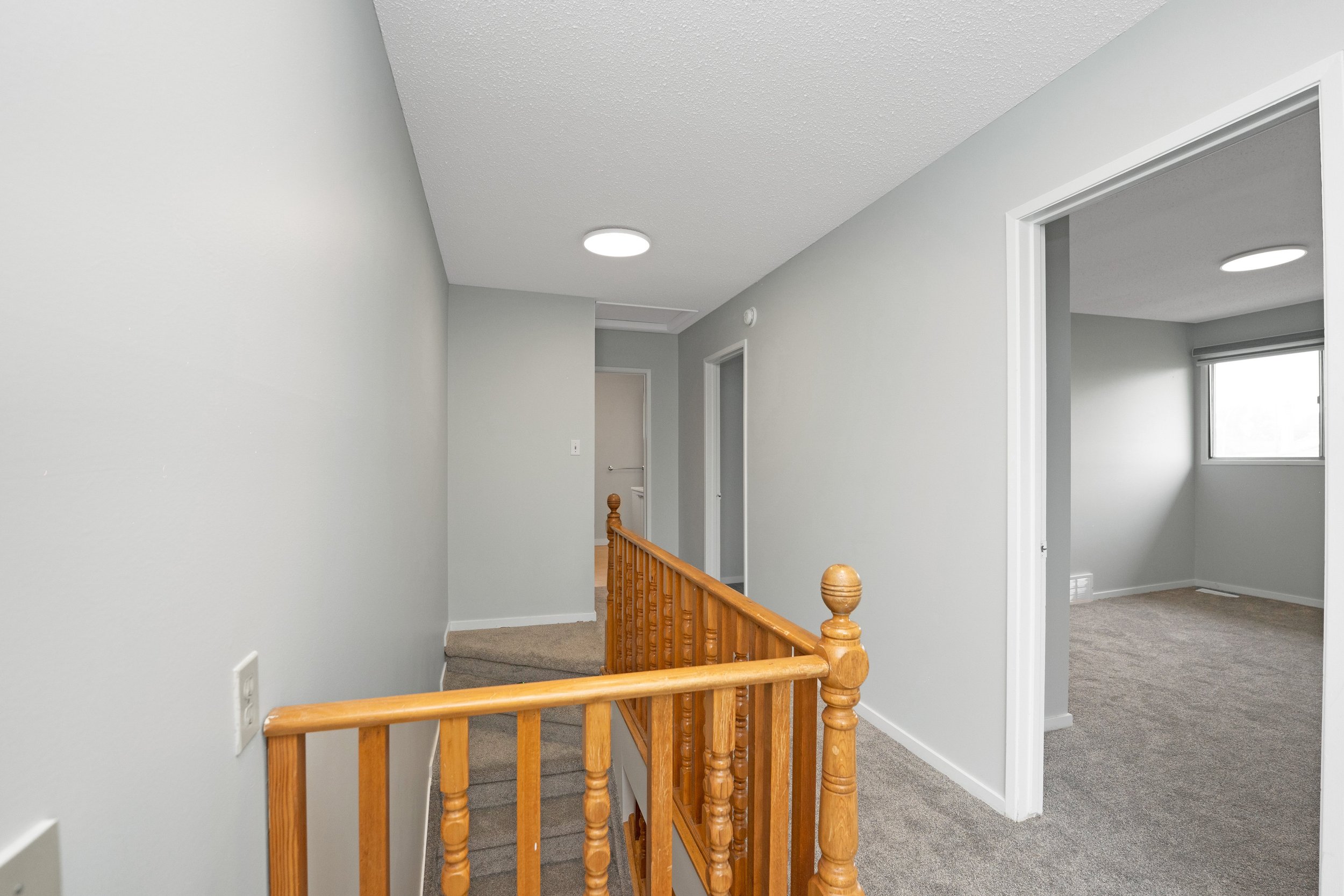 Interior view of a hallway with gray walls, wooden handrail, carpeted floors, and a room with a window on the right.