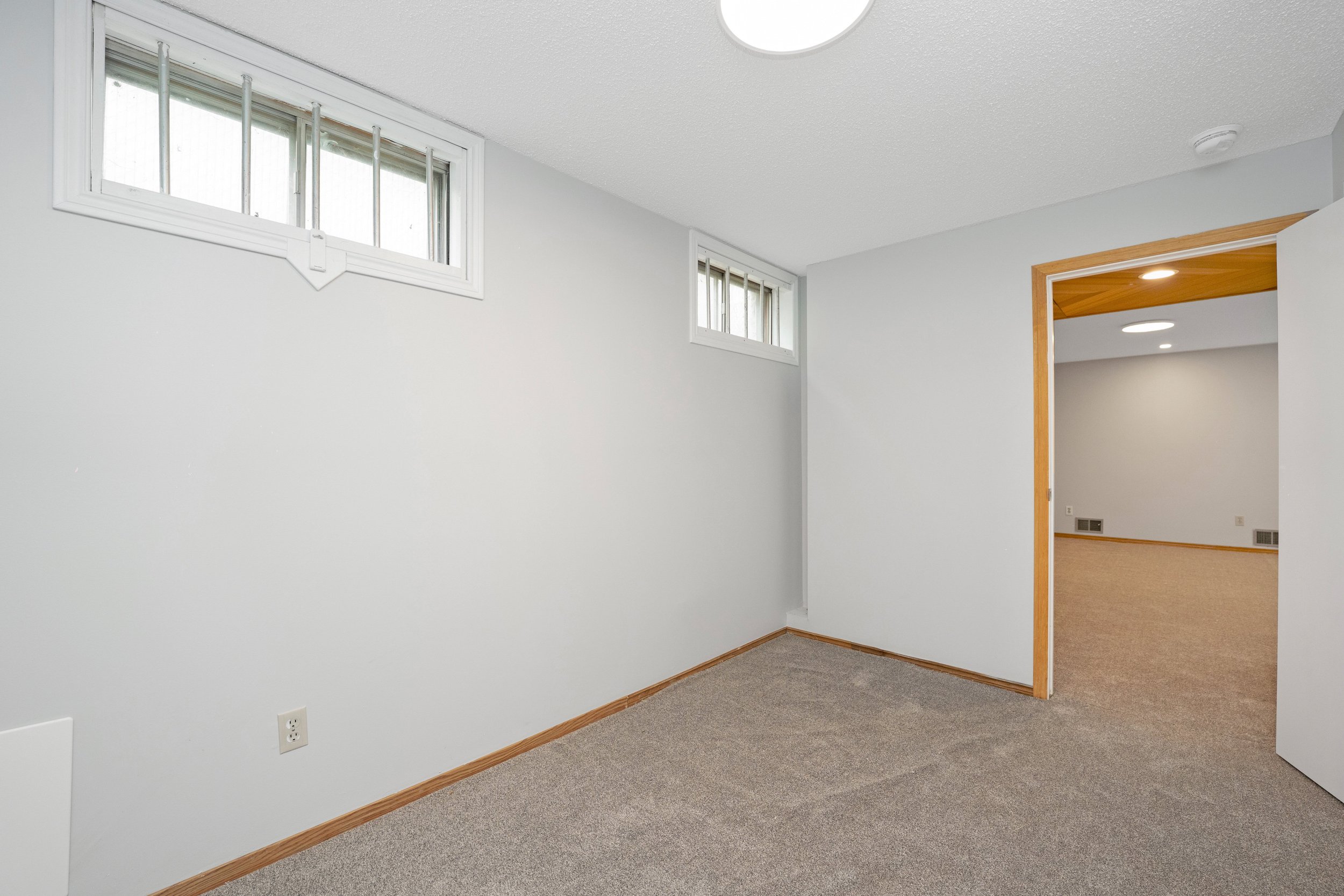 Empty room with white walls, gray carpet, and small rectangular windows with bars, leading into another room with beige carpet and wall vents.
