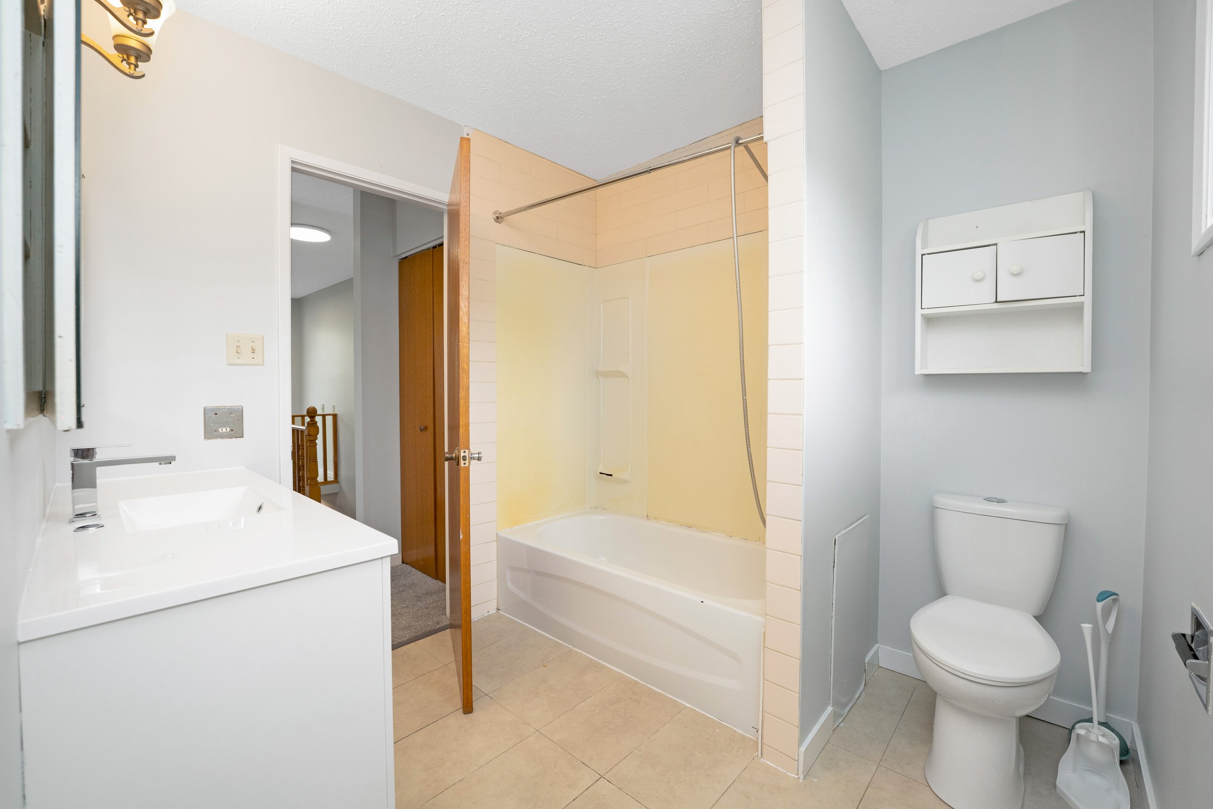 Bathroom with a white vanity and sink, yellow and white bathtub with shower, toilet with a white shelf above, light-colored tiled floor, light gray walls, small window, and partially open wooden door leading to a hallway.