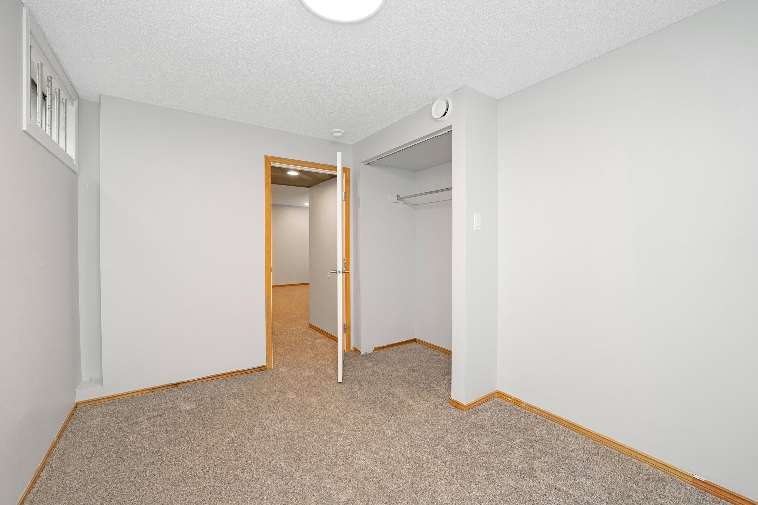 Empty room with beige carpet, white walls, wooden trim, an open closet with a metal rod, small window, and an open door leading to a hallway.
