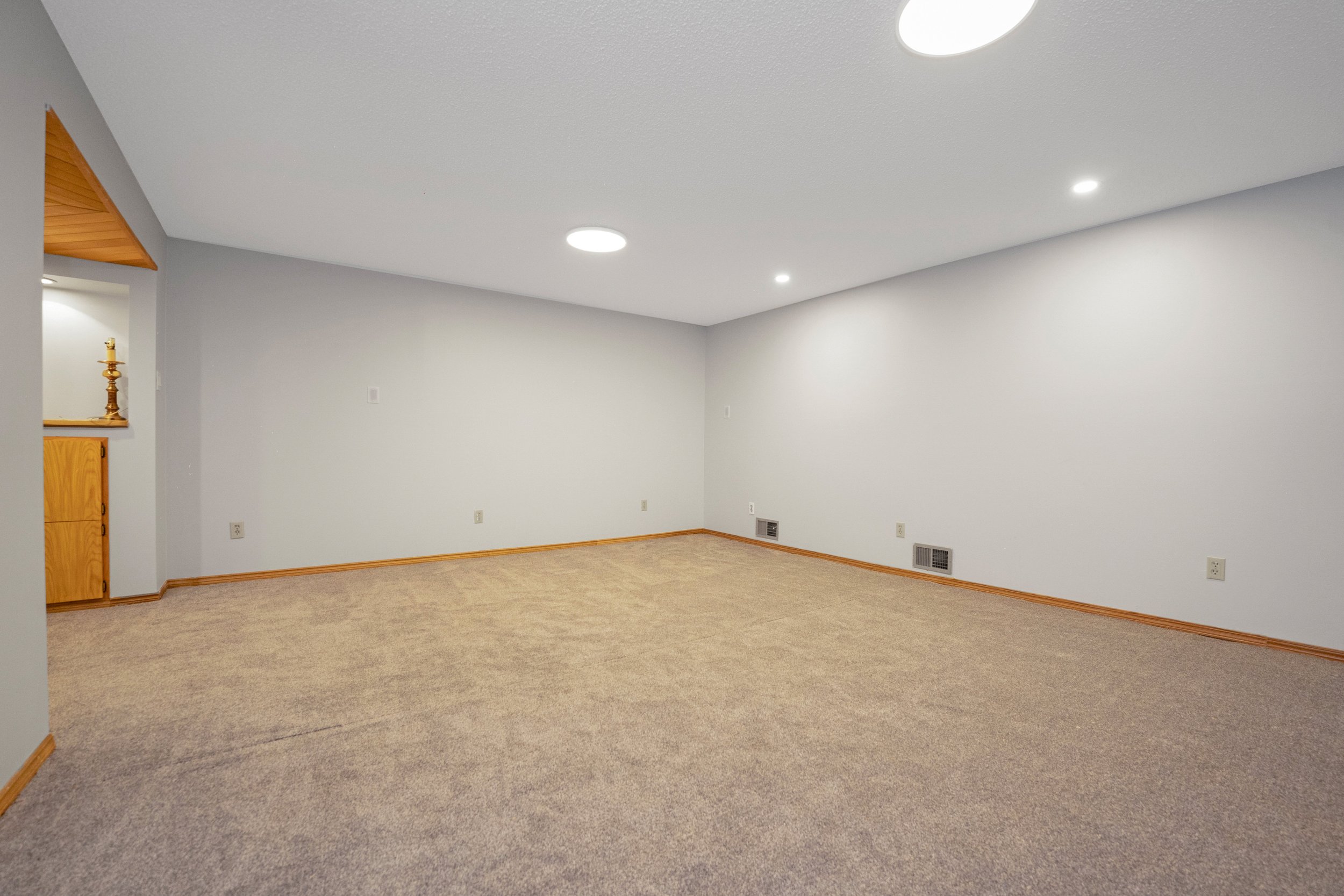 Empty living room with beige carpet, plain white walls, ceiling lights, and wooden baseboards.