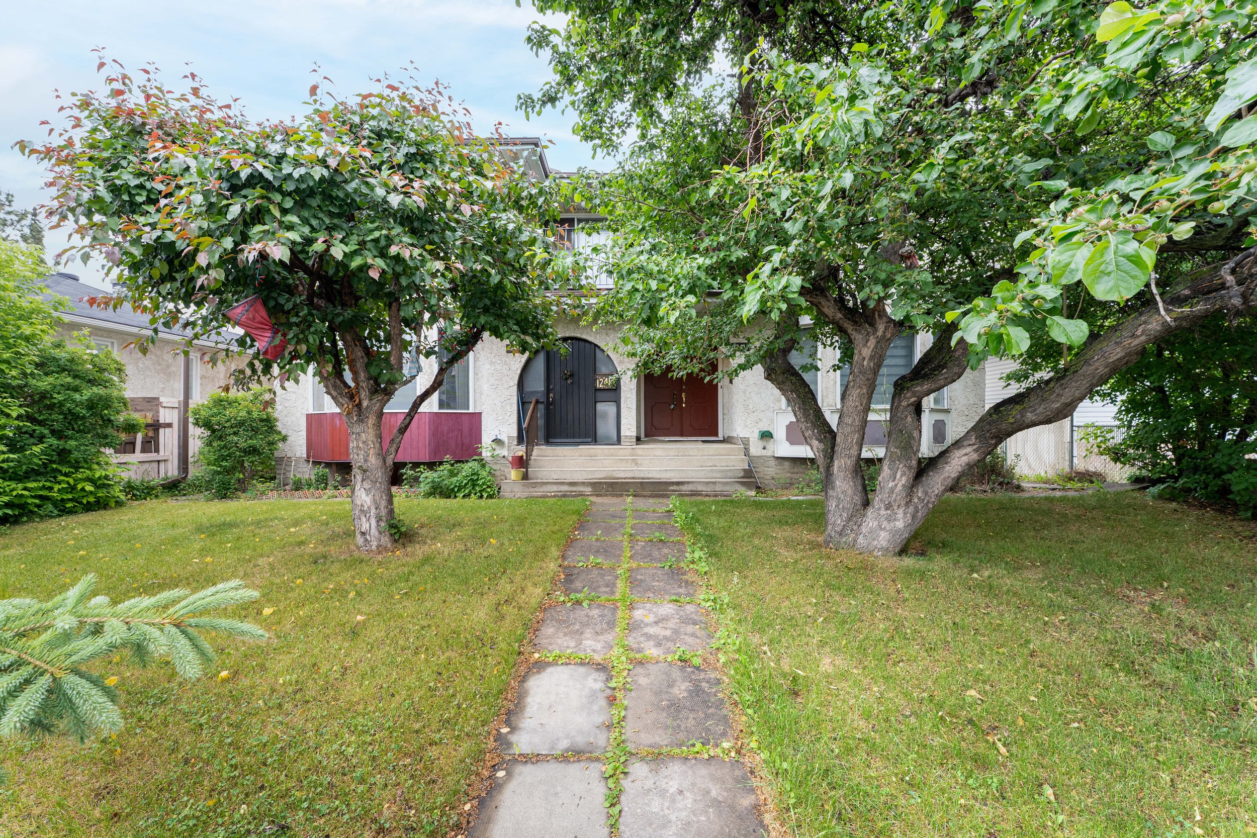 Front yard with a stone pathway leading to a house entrance, flanked by two large leafy trees.