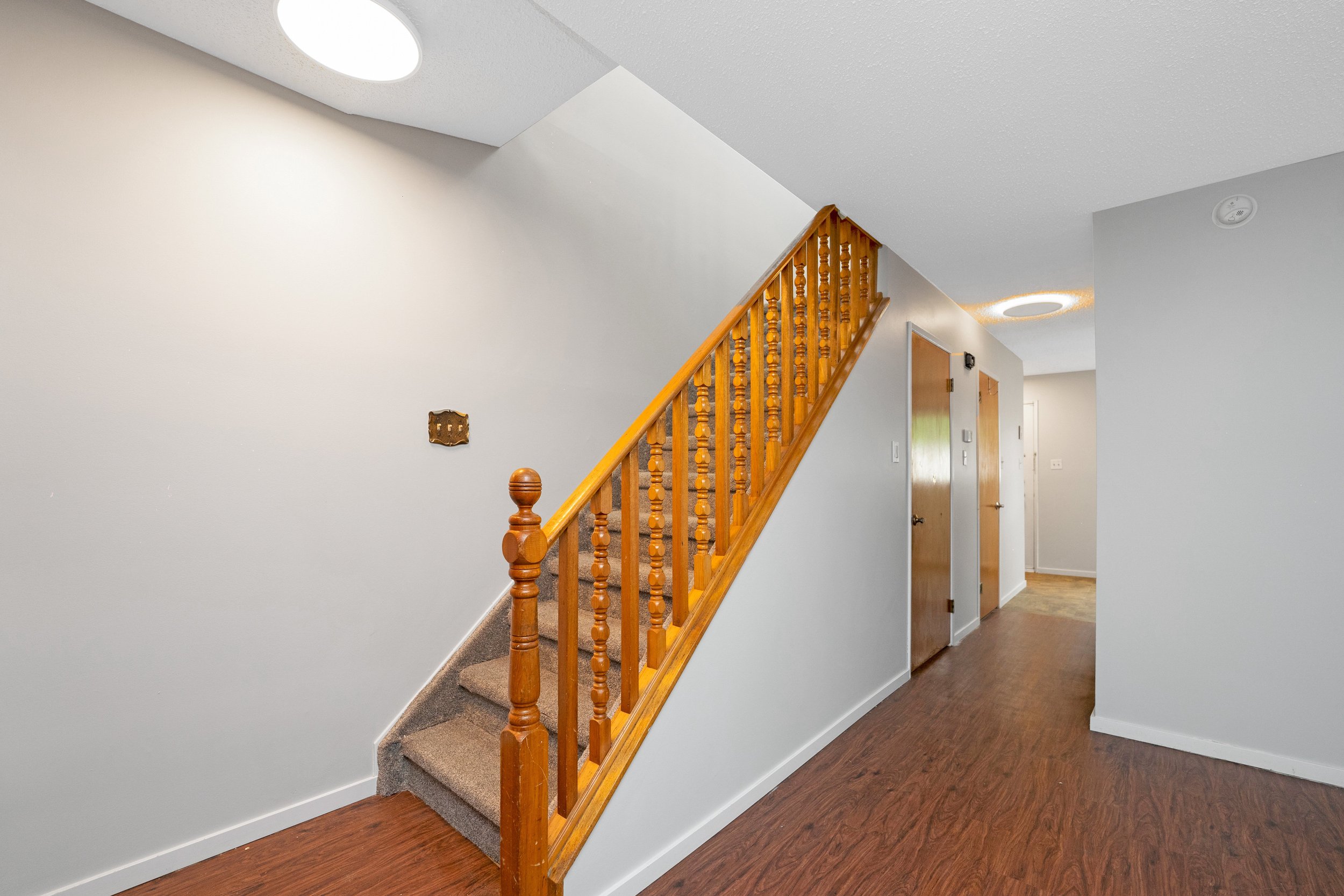 Interior view of a home with a staircase with wooden railings, beige carpet, white walls, and hardwood flooring.