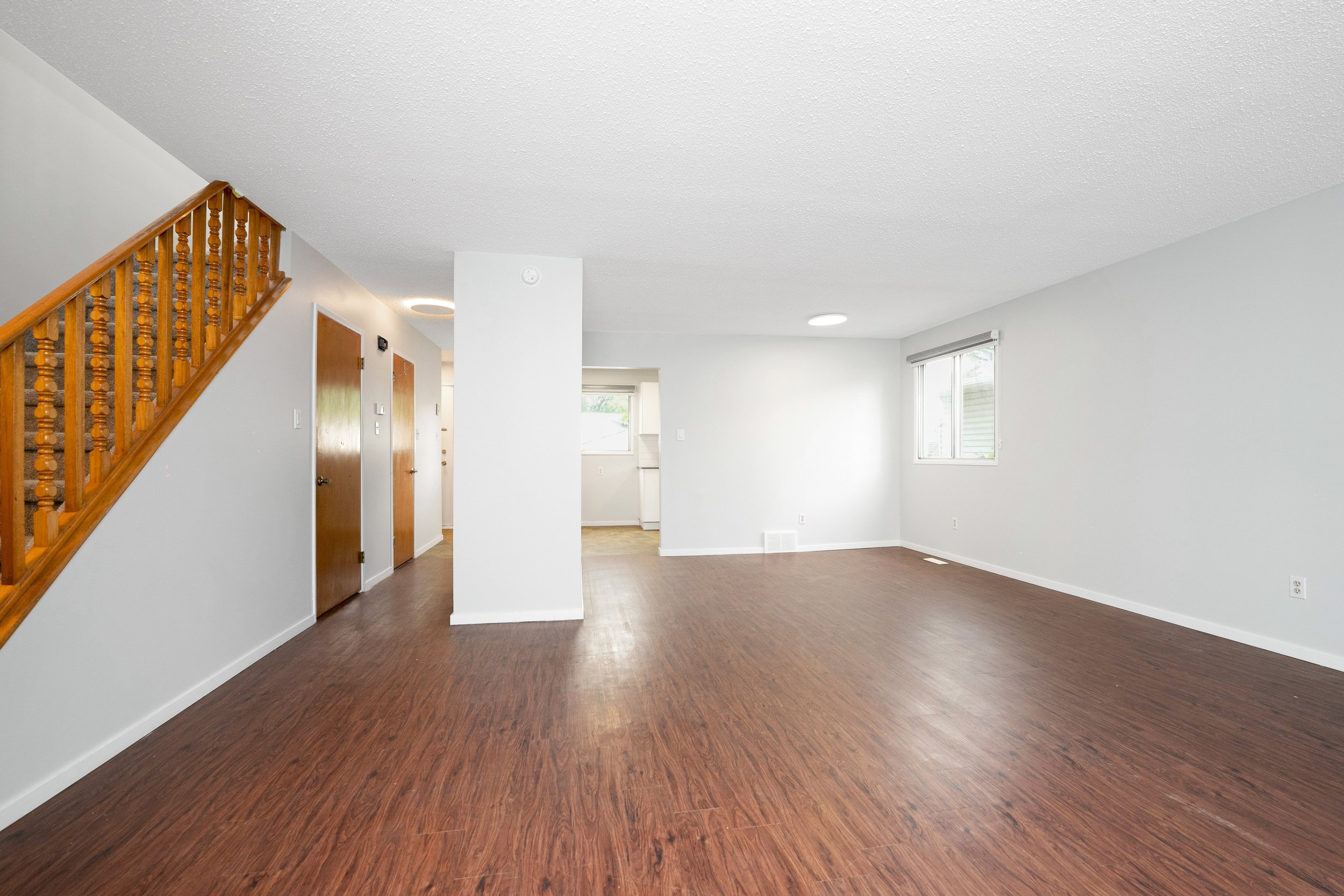 Empty living room with hardwood flooring, white walls, and two windows, with a wooden staircase on the left side leading upstairs.