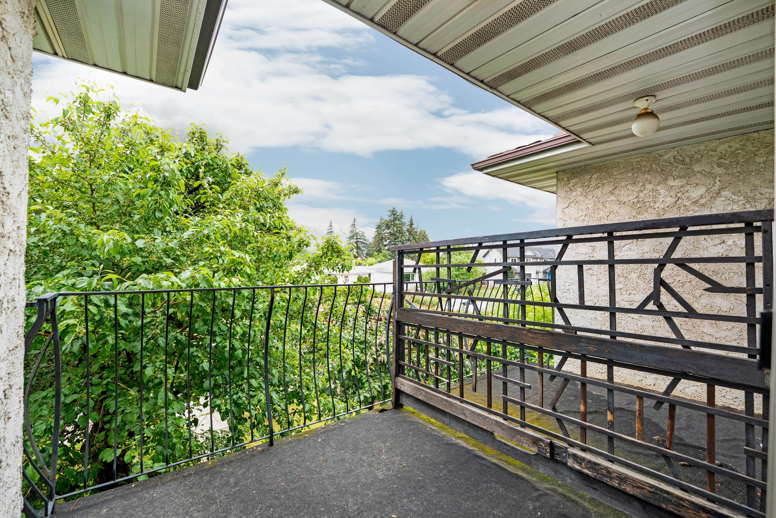 View from a balcony showing a black metal railing, a green leafy tree, a neighboring house, and a partly cloudy sky.