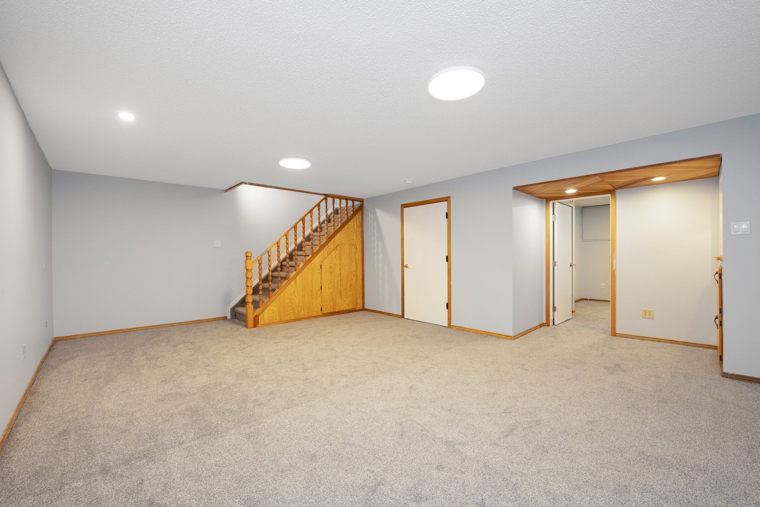 Empty living room with beige carpet, light gray walls, wooden trim, and built-in niches with wood paneling; staircase with wooden railing to the left.