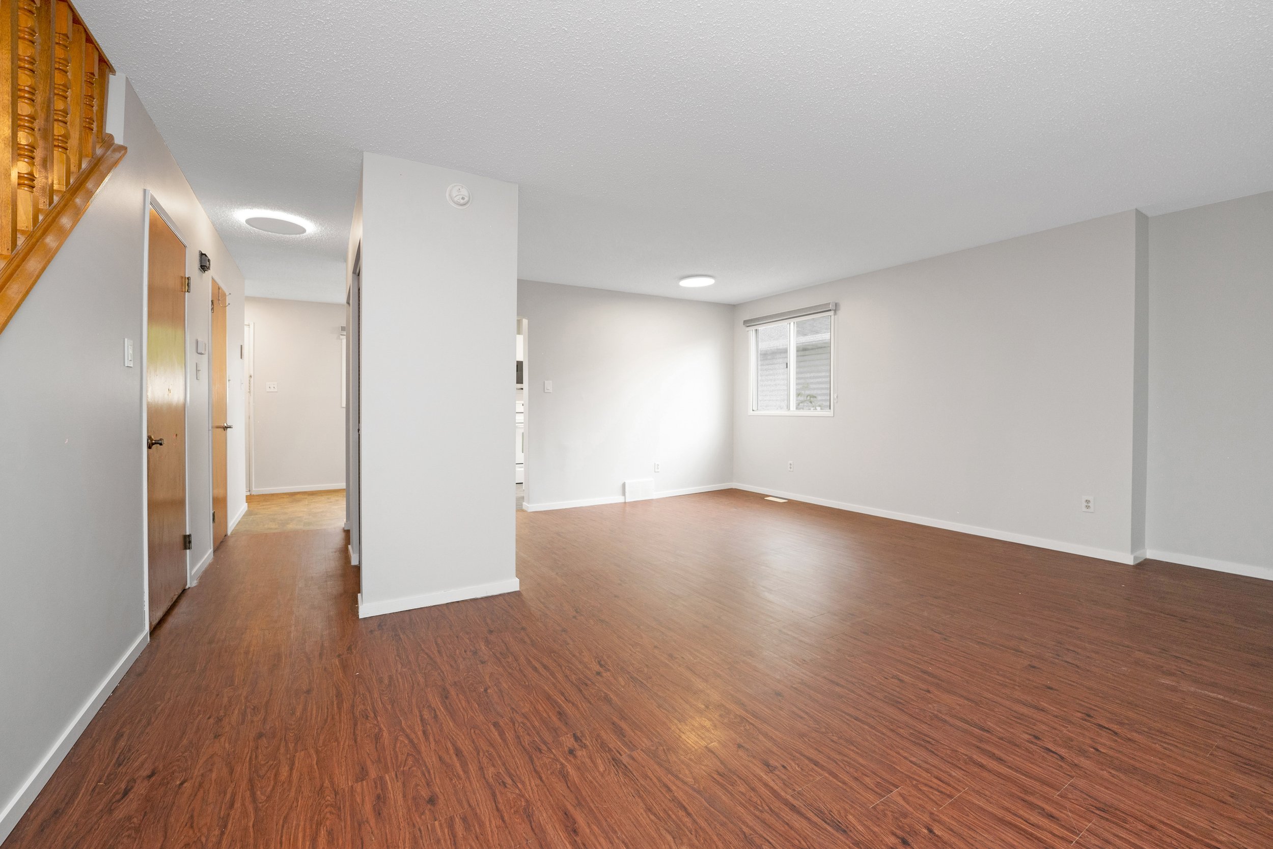 Empty living room with white walls, wood flooring, window, and staircase.