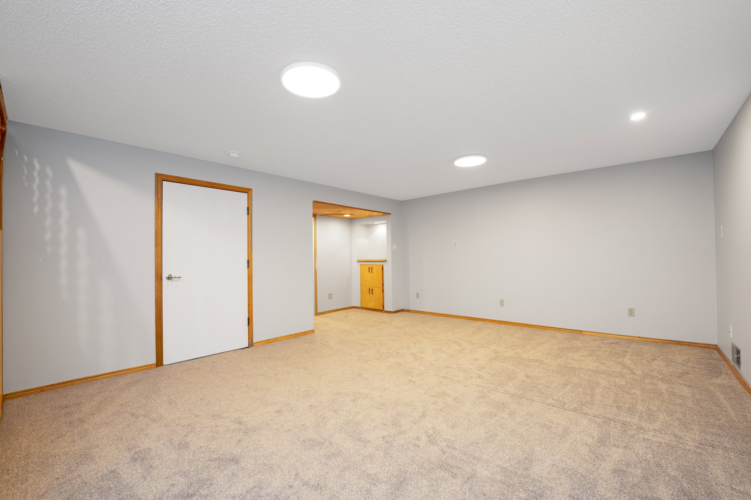Empty room with beige carpet, white walls, wood trim, and ceiling lights. There are two closed white doors with wood frames and a small built-in wooden cabinet in the corner.