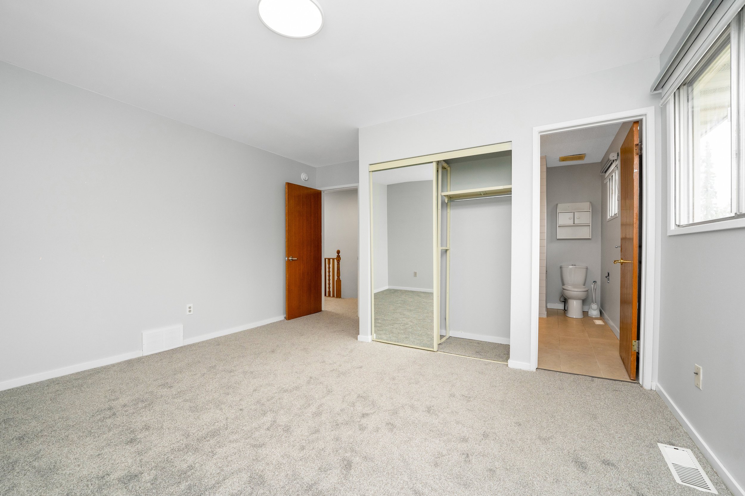 Empty bedroom with beige carpet, white walls, and a ceiling light. There is an open closet with a mirror sliding door. An open door reveals a bathroom with a toilet and small window. A second door leads to a hallway with a staircase.