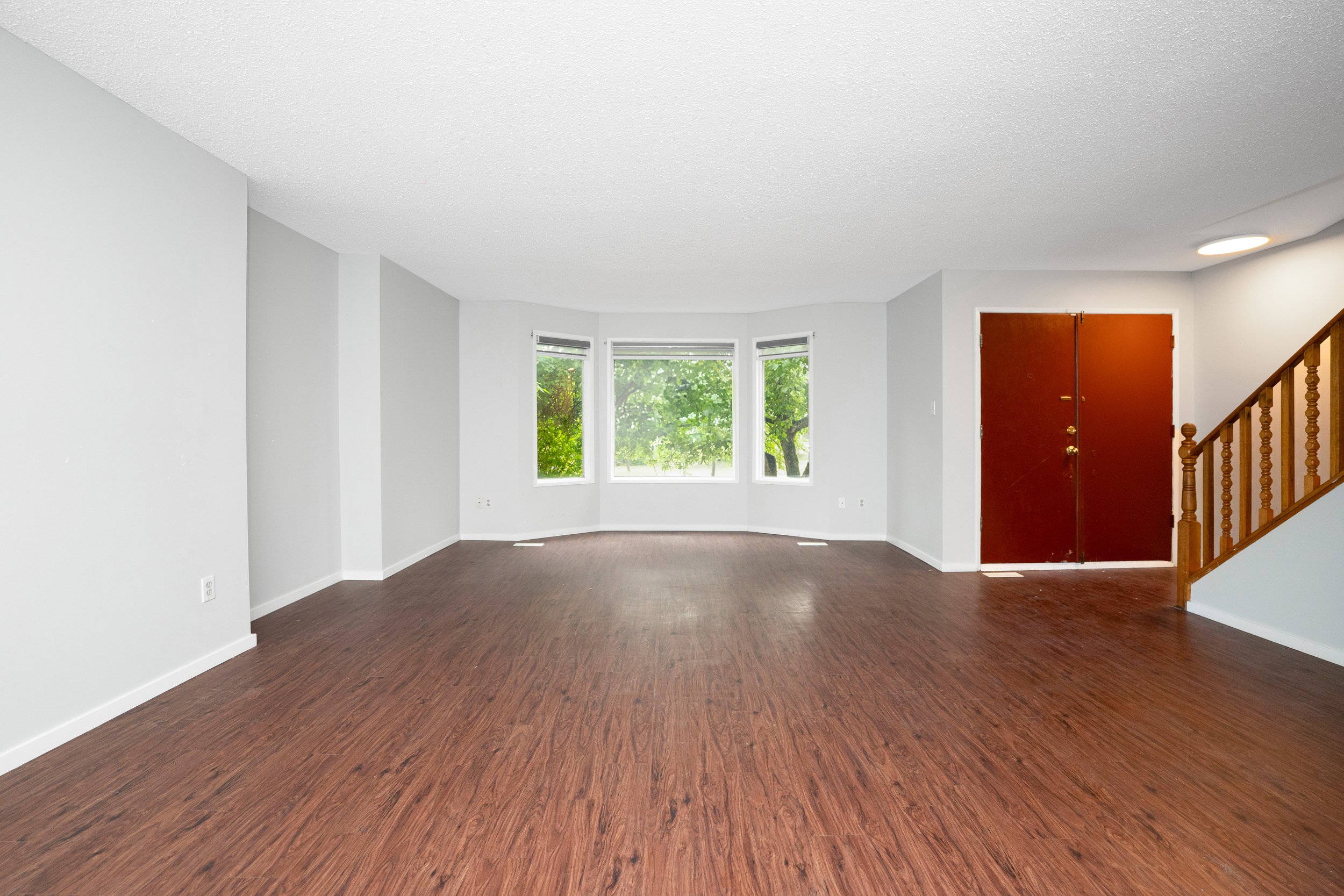 Empty living room with white walls, hardwood floors, a large window bay with greenery outside, a wooden staircase, and a reddish-brown front door.