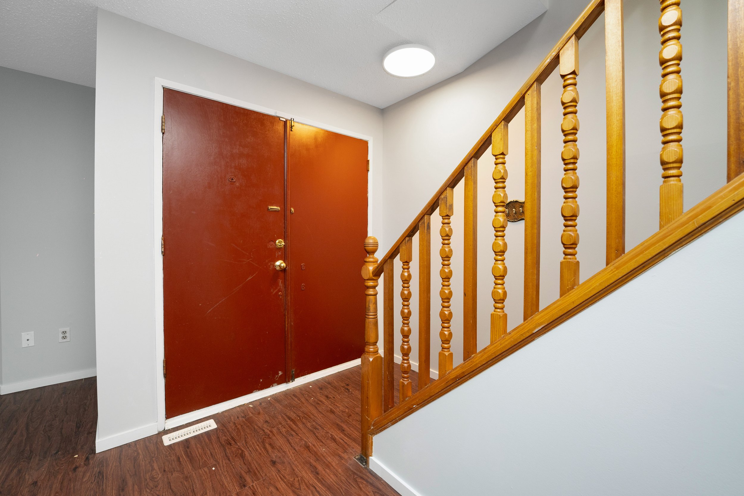 Interior of an entryway with a red front door, wooden staircase railing, gray walls, and wood flooring.