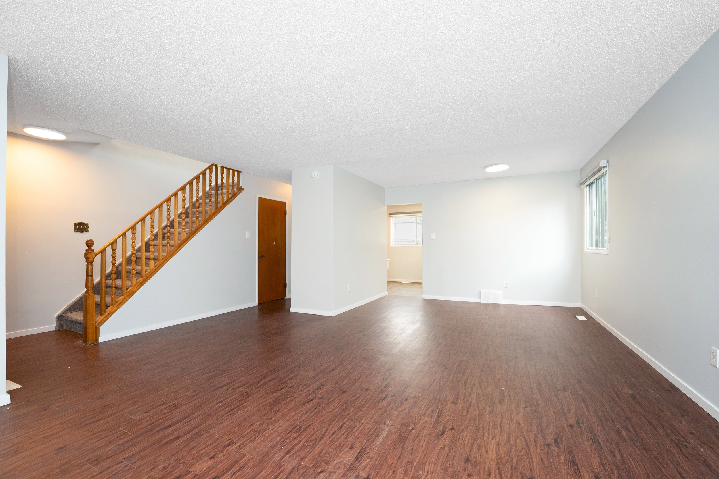Empty living room with hardwood floors, white walls, a staircase with wooden railing, and windows letting in natural light.